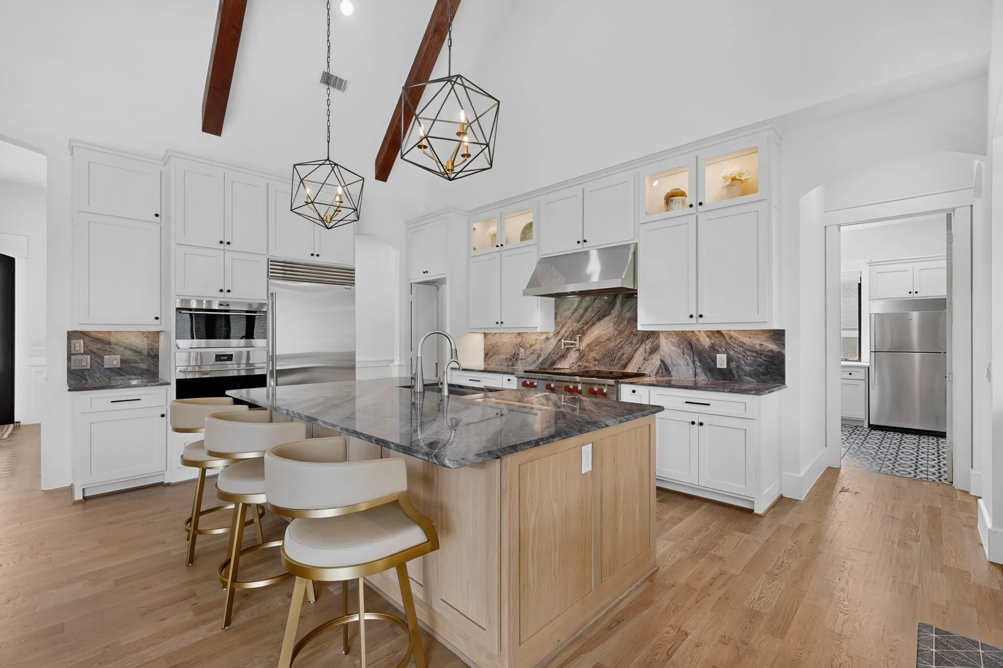 Vaulted ceiling kitchen featuring exposed beams, light wood-finish flooring, a large island with a quartzite countertop, white cabinetry with display uppers, and stainless steel Wolf and SubZero appliances