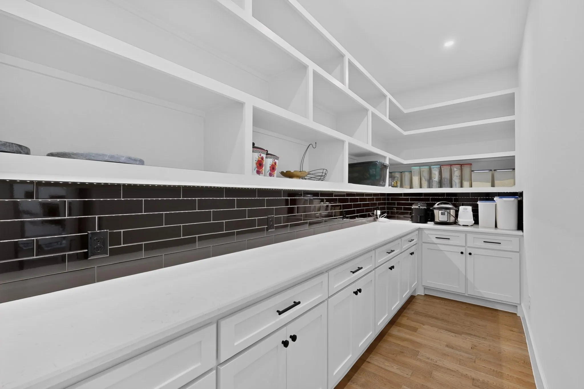 Hidden doors hide the spacious pantry featuring extensive open shelving, white cabinetry, integrated countertop, dark subway tile backsplash, and wood-finish flooring behind the kitchen