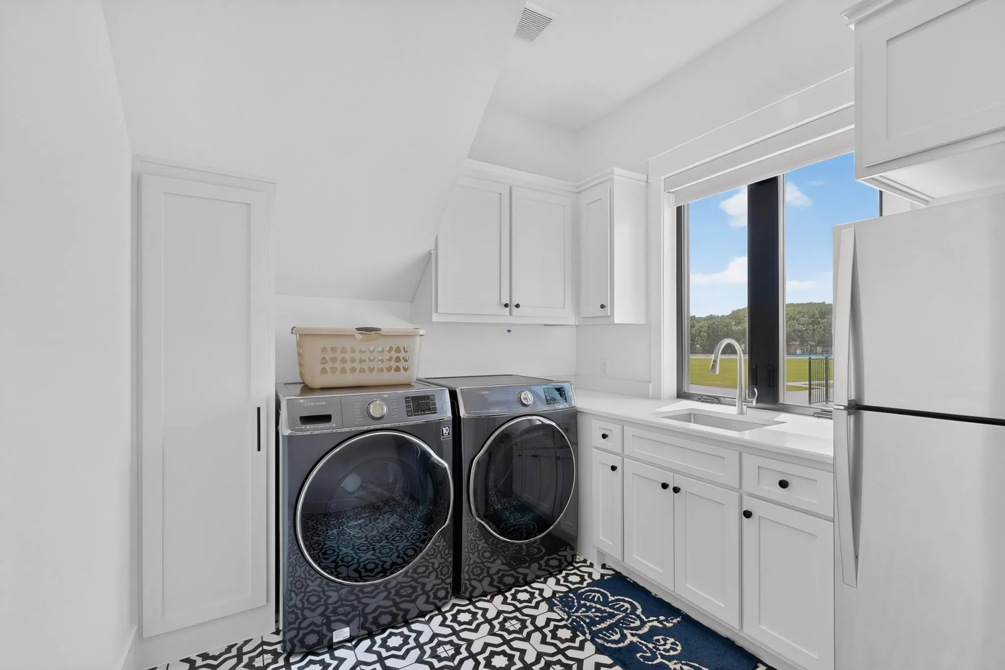 Functional laundry room featuring white cabinetry, a sink with a gooseneck faucet, light-toned countertops, and patterned floor tiles