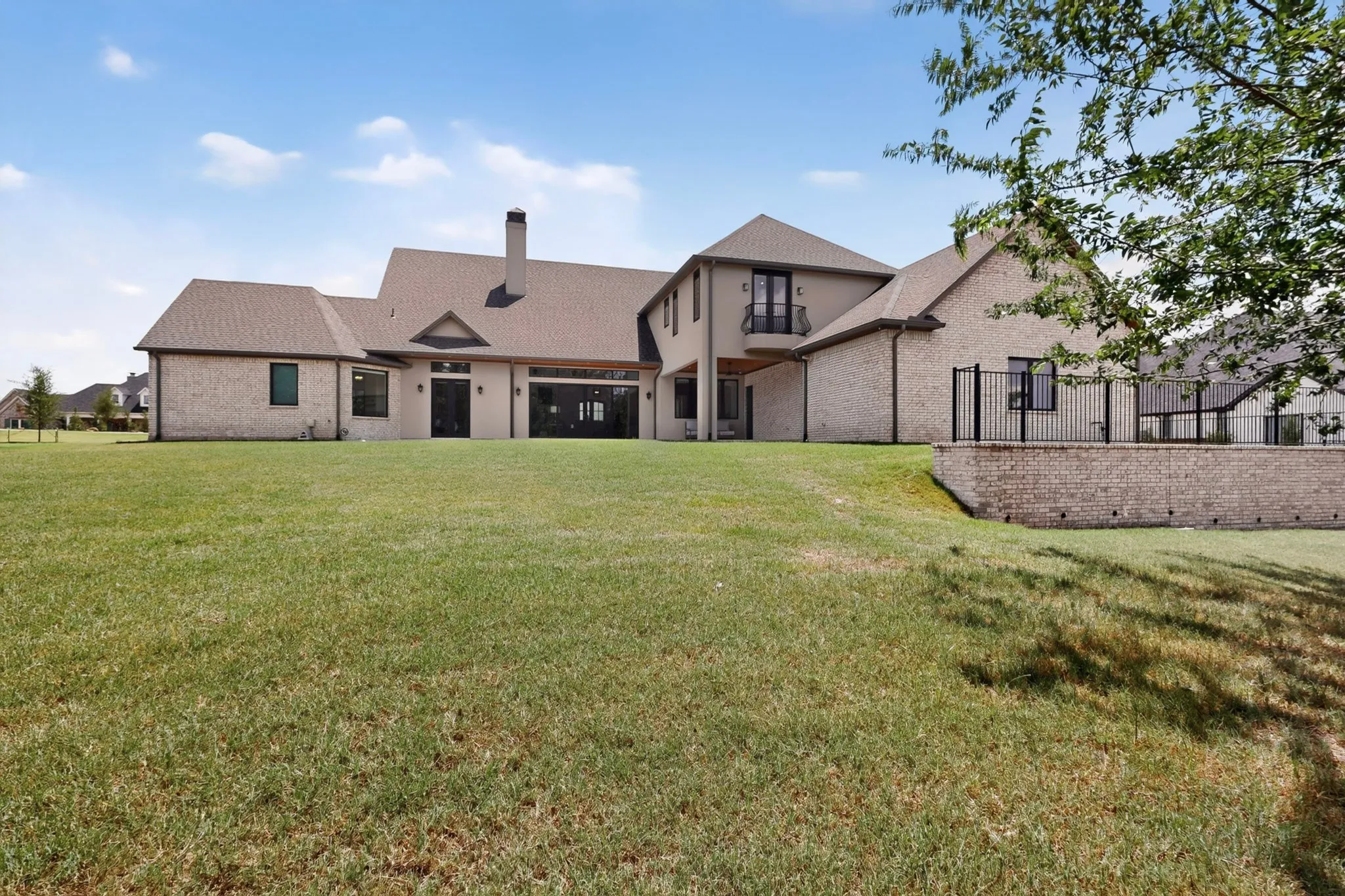 Expansive backyard with a manicured lawn, brick and stucco exterior, large sliding glass doors, a second-story balcony with wrought iron railing, and a retaining wall with fencing