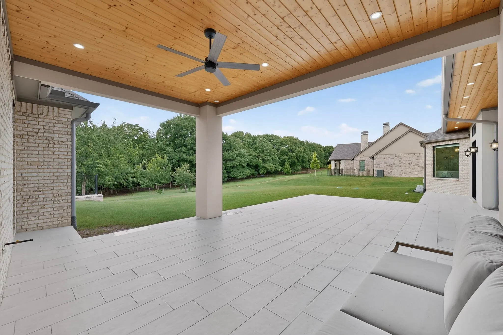 Covered outdoor patio featuring light-toned rectangular tile flooring with views to the large backyard framed with huge oak trees