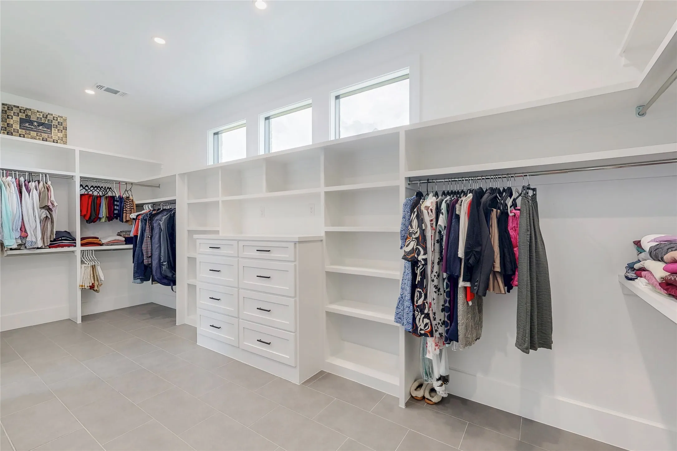 Spacious Primary custom closet featuring large windows for natural light, extensive built-in shelving, multiple hanging rods, built-in cabinet hutch storage, and light-tone large format tile flooring