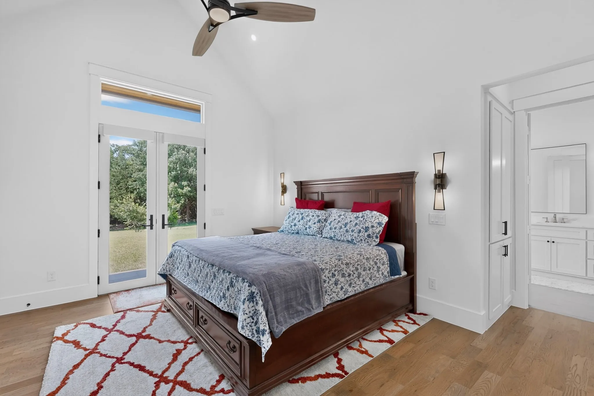 Spacious Primary Bedroom featuring vaulted ceilings, wood-finish flooring, and French doors with transom window and sconce lighting frame the bed area.