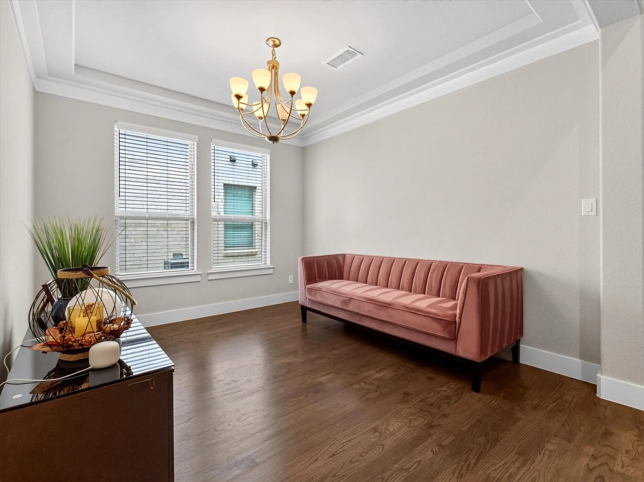 Formal room with rich wood-finish flooring, featuring a tray ceiling with crown molding, a brass-tone chandelier, and two large windows with blinds