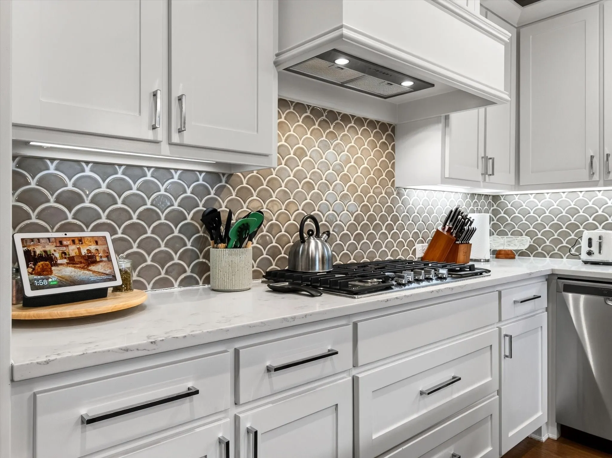 White cabinetry with contemporary hardware, scalopped tile backsplash, under-cabinet lighting, a built-in cooktop, and a stainless steel dishwasher