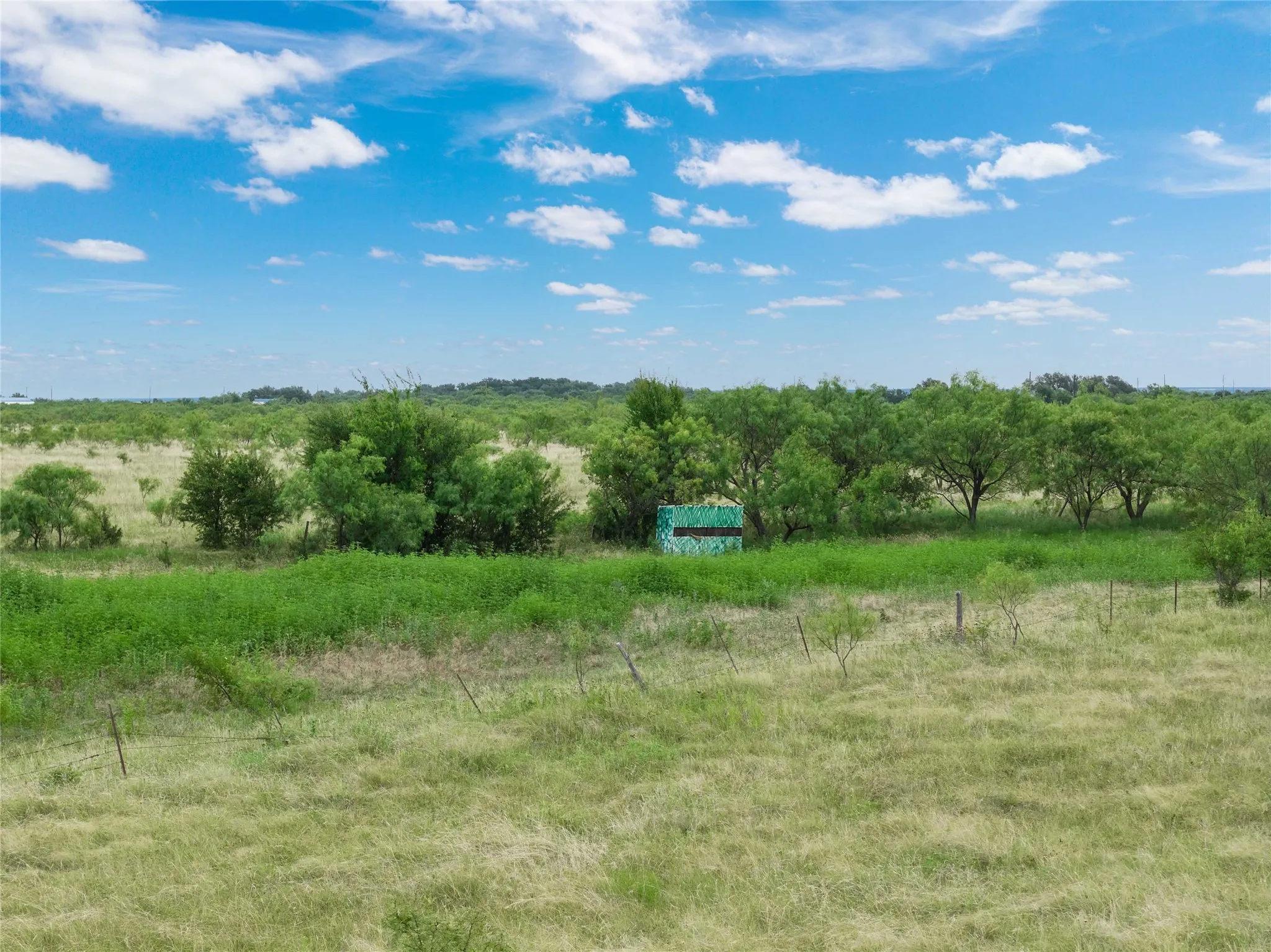 Expansive green landscape featuring varied vegetation, a wire fence line, and a small structure with a camouflage pattern