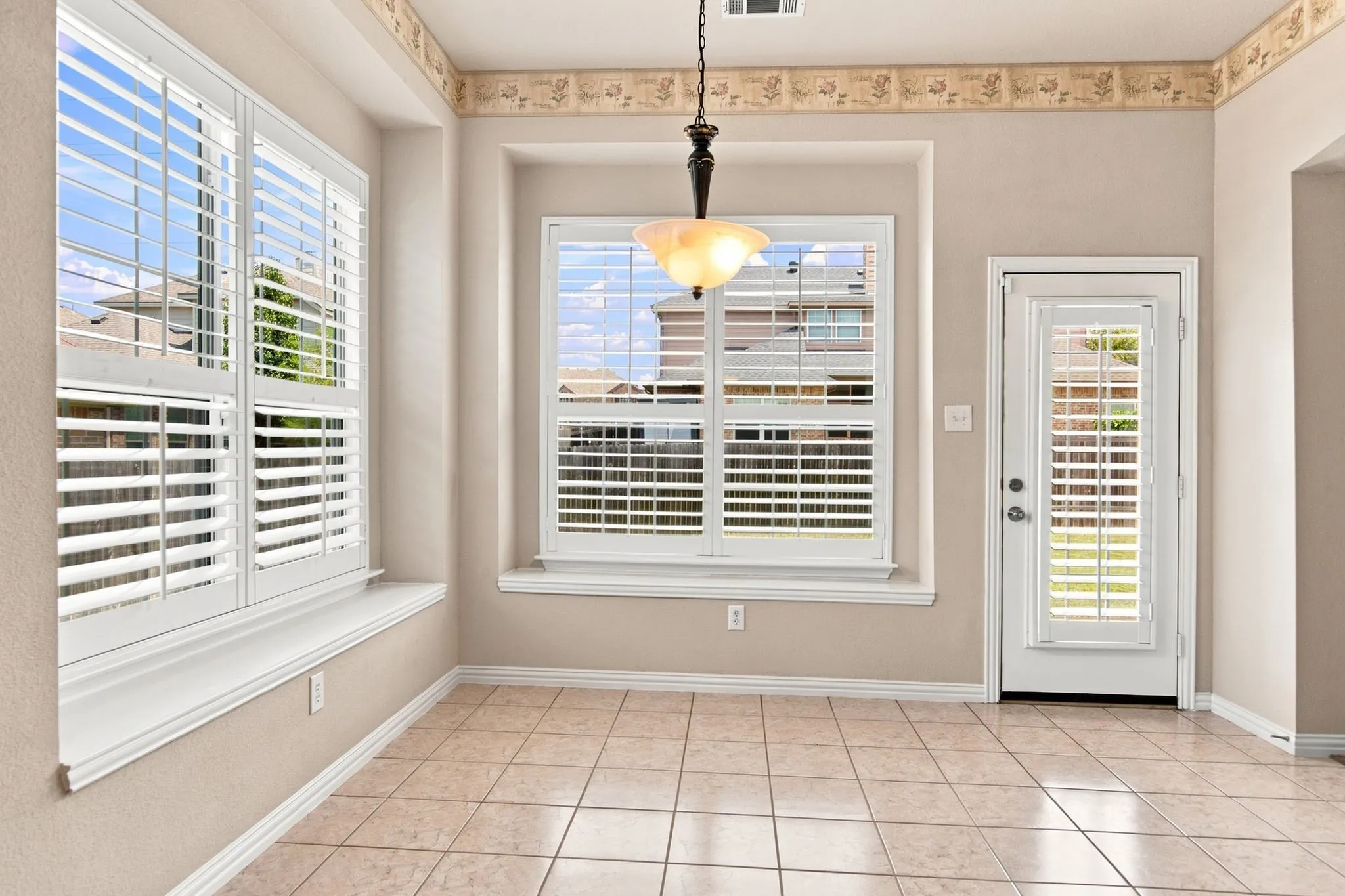 Tile flooring, multiple windows with plantation shutters, ceiling-mounted light fixture, and a glass-paneled door