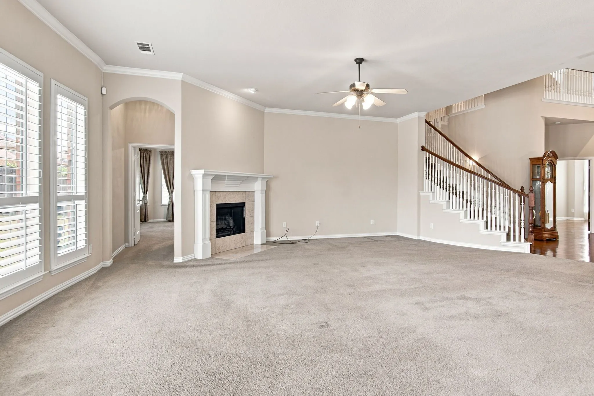 Spacious living area featuring a fireplace with a mantel, multiple windows with plantation shutters, a ceiling fan, and a grand staircase with wood railings