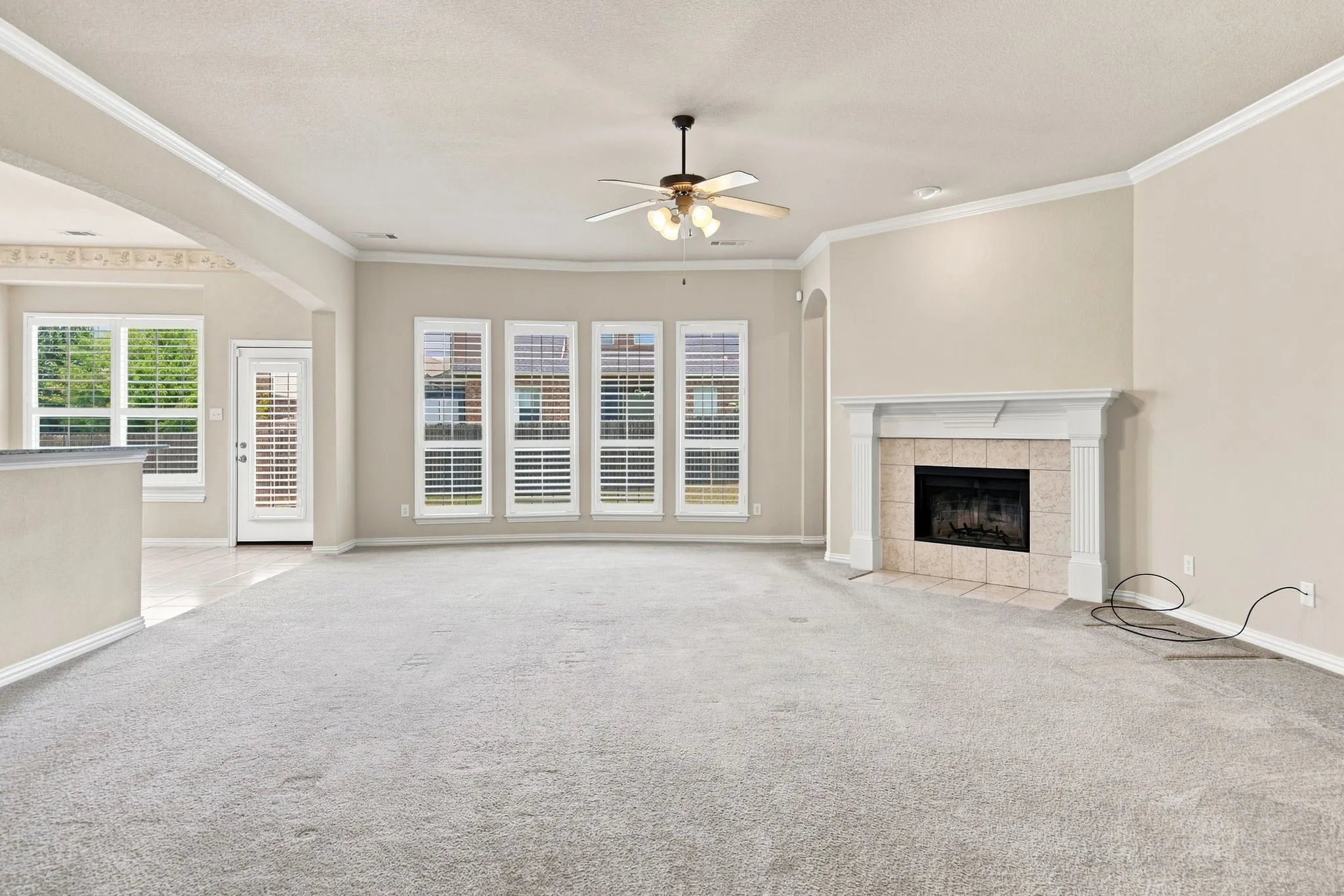 Spacious living area featuring a fireplace with a stone surround, multiple windows with plantation shutters, a ceiling fan, and crown molding
