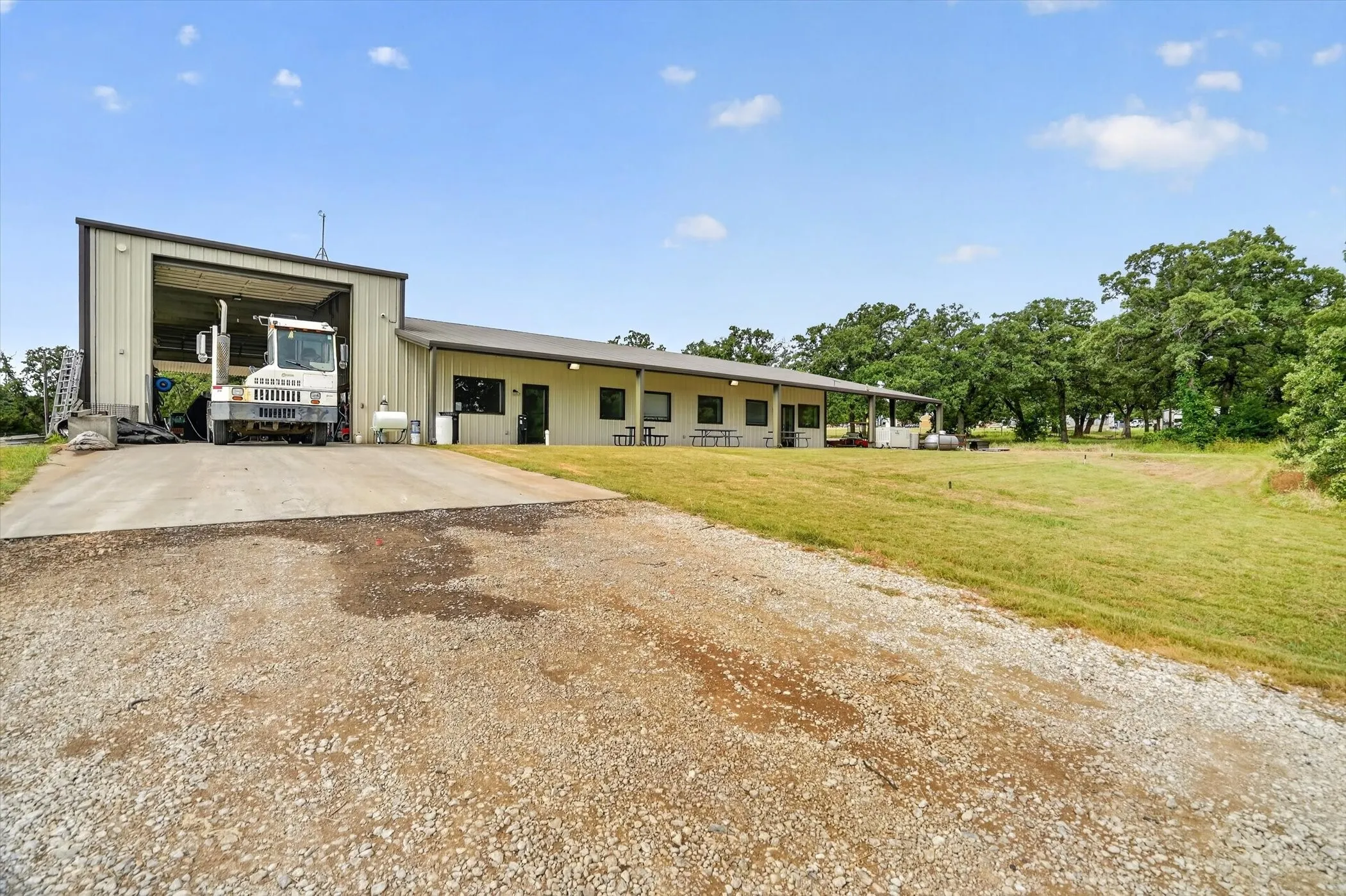 Metal-clad building featuring a large open bay, concrete drive, covered patio, and surrounding expansive green space with mature trees