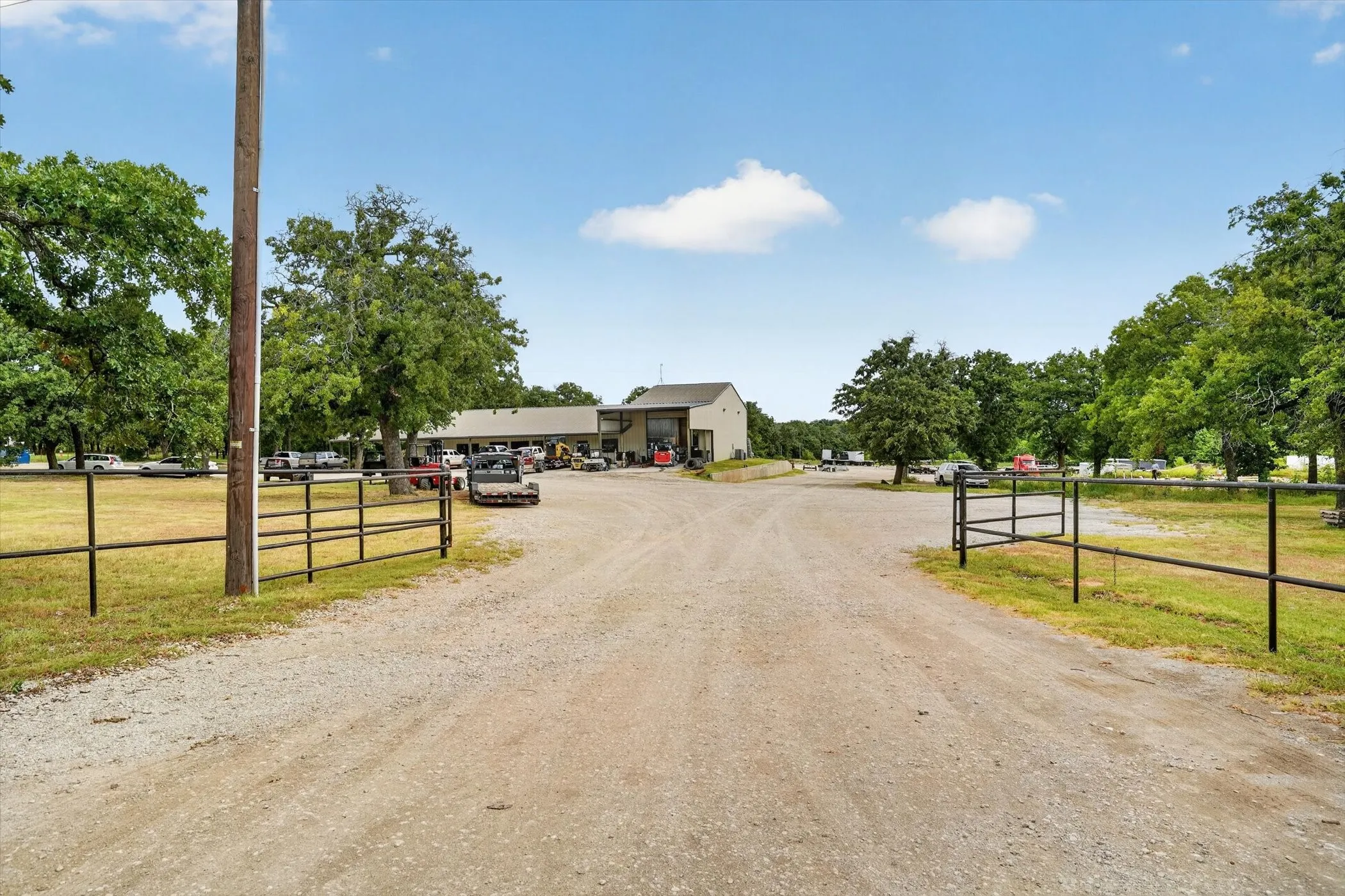 Gravel driveway entry with double metal gates, mature trees, and multiple outbuildings