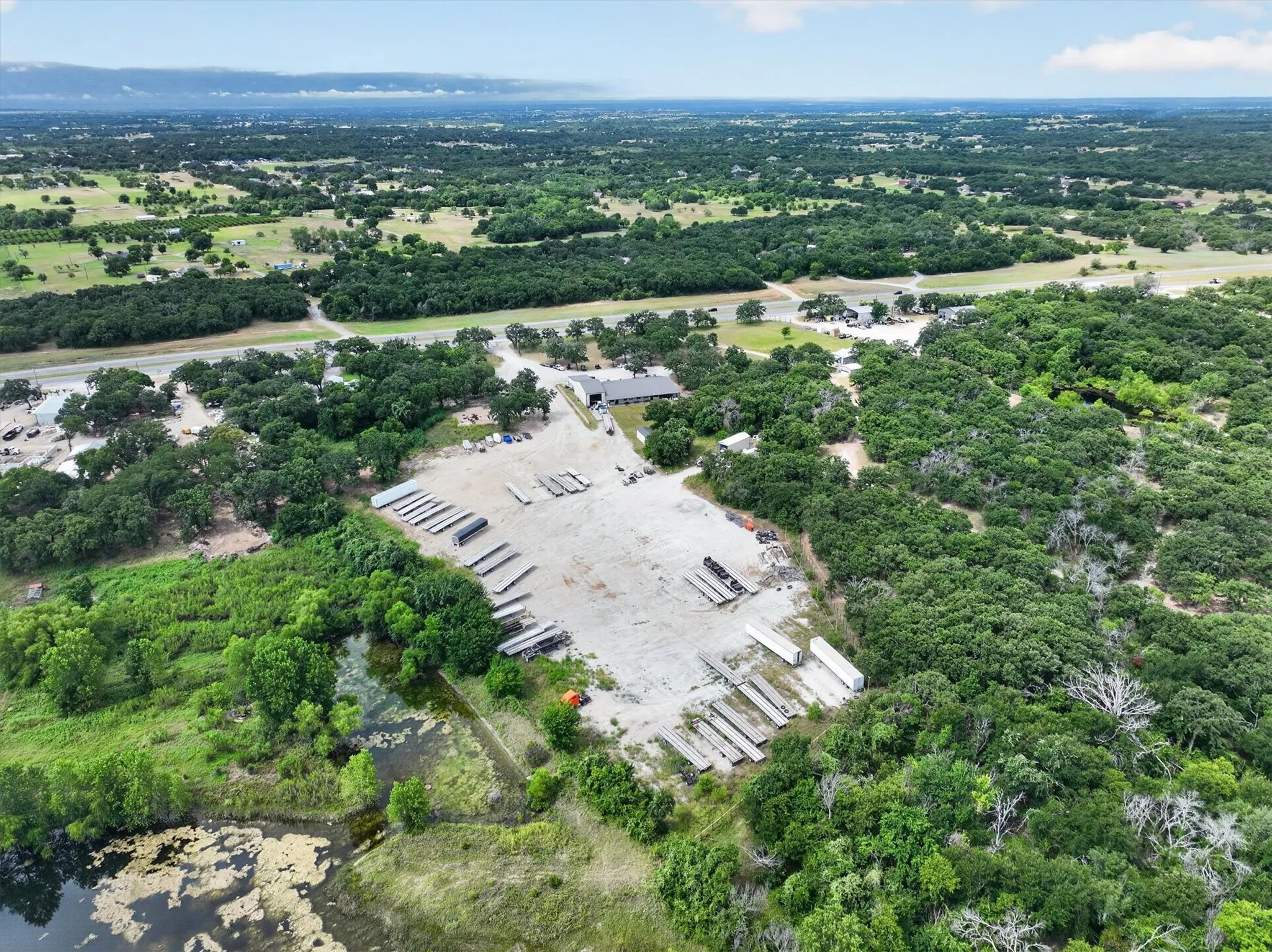 Expansive land parcel featuring a large gravel lot, dense tree coverage, a body of water, a single-story building, and direct road access