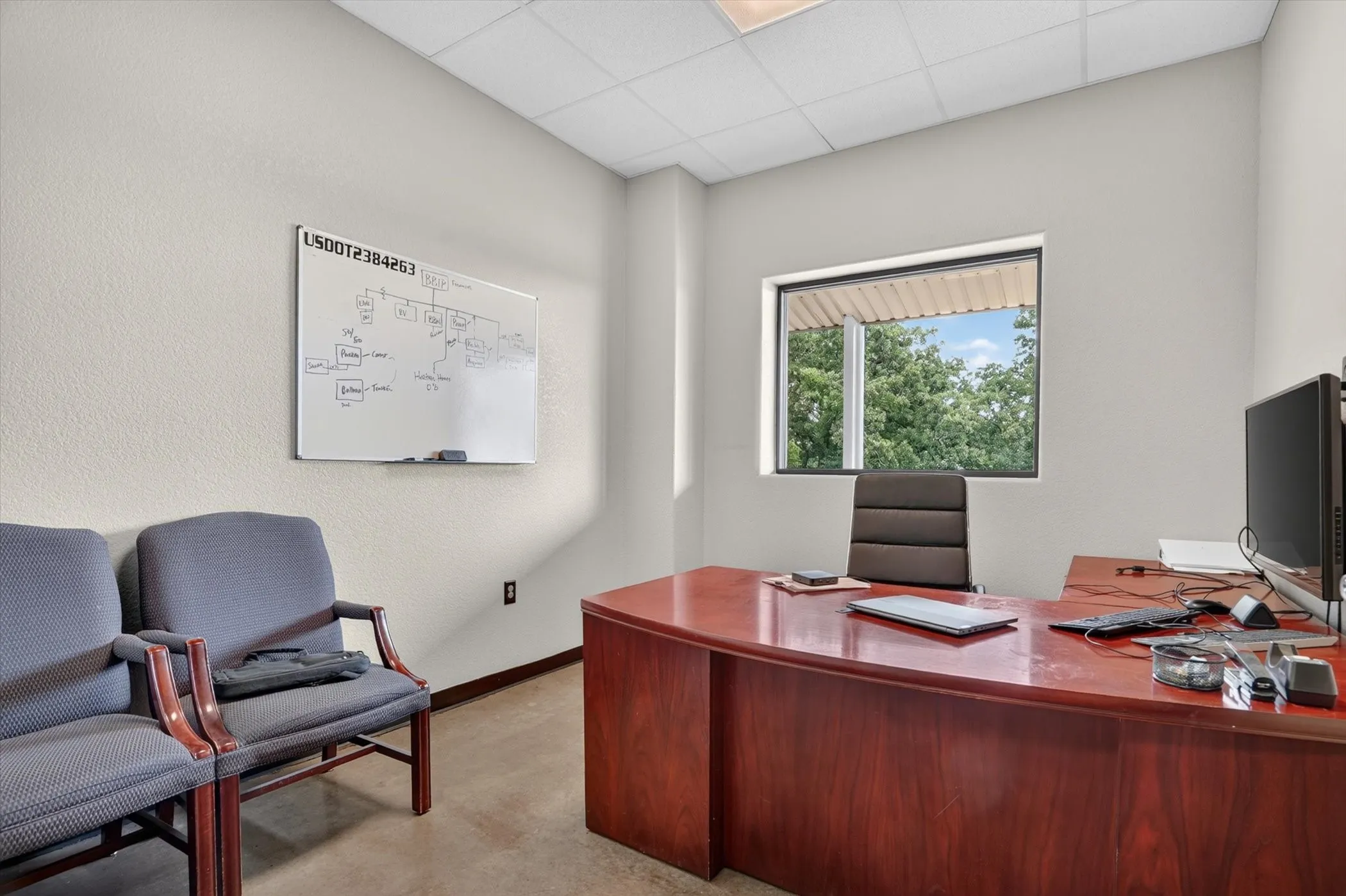 Office space featuring a wood-finish desk, ample natural light from the window, textured light-colored walls, and a dropped ceiling with integrated lighting