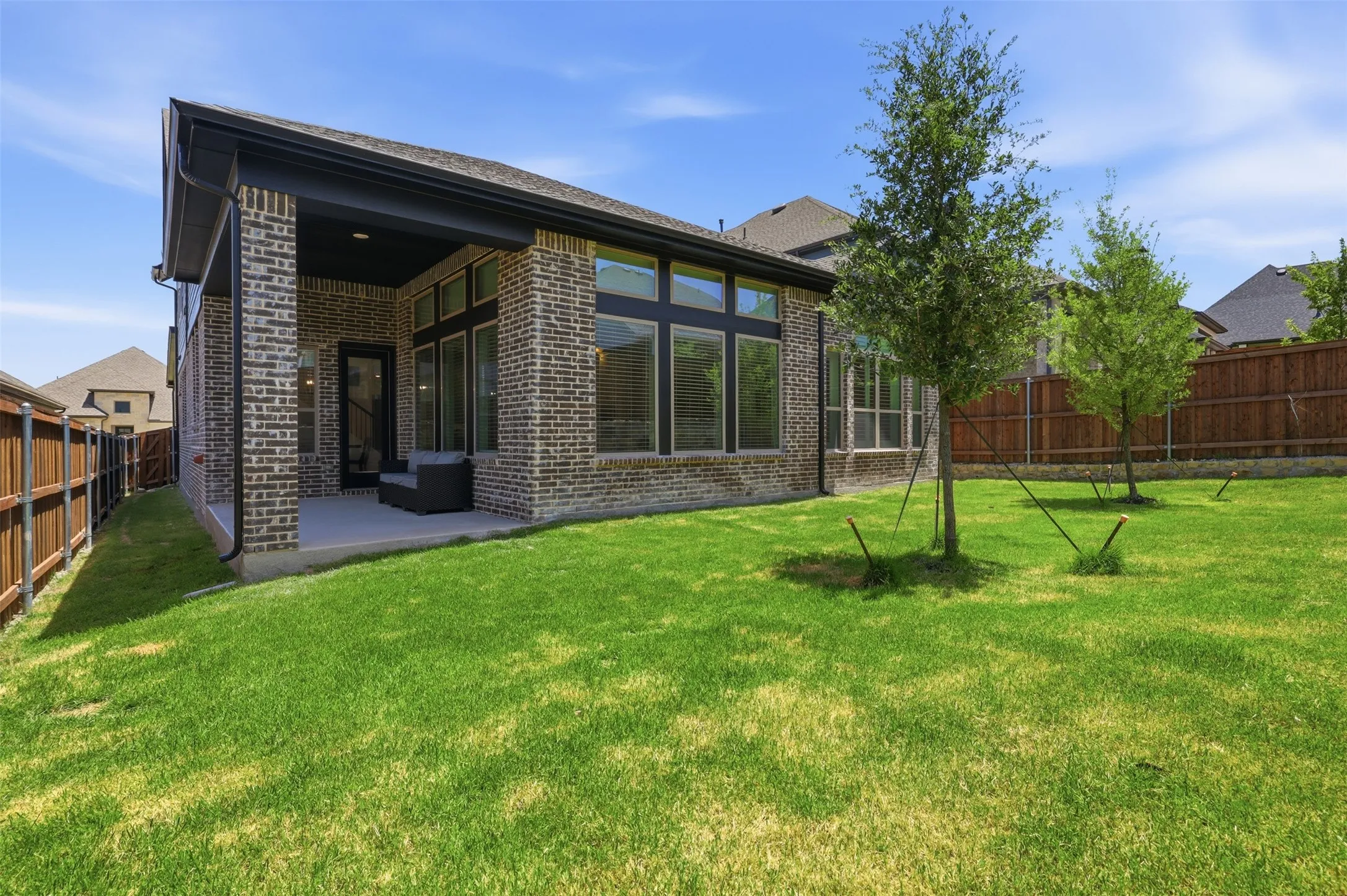Expansive rear lawn featuring a covered patio, brick exterior, dark frame windows, and young trees