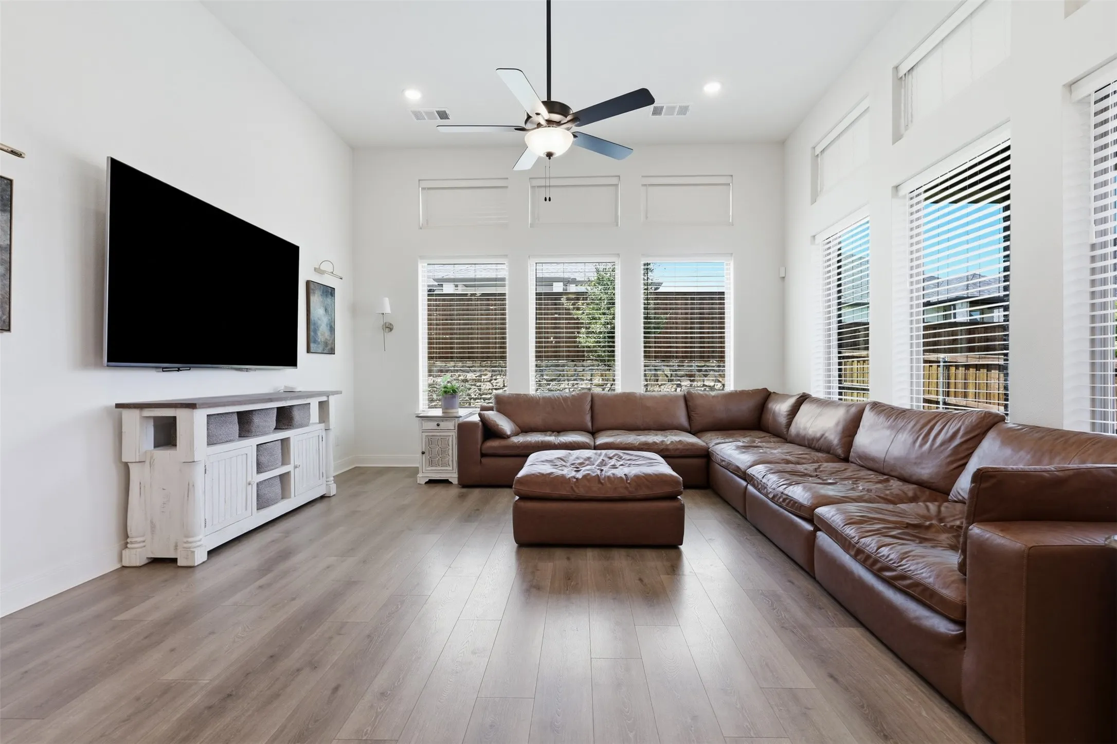 Spacious living area featuring high ceilings, wood-finish flooring, a ceiling fan with integrated lighting, and multiple windows with blinds