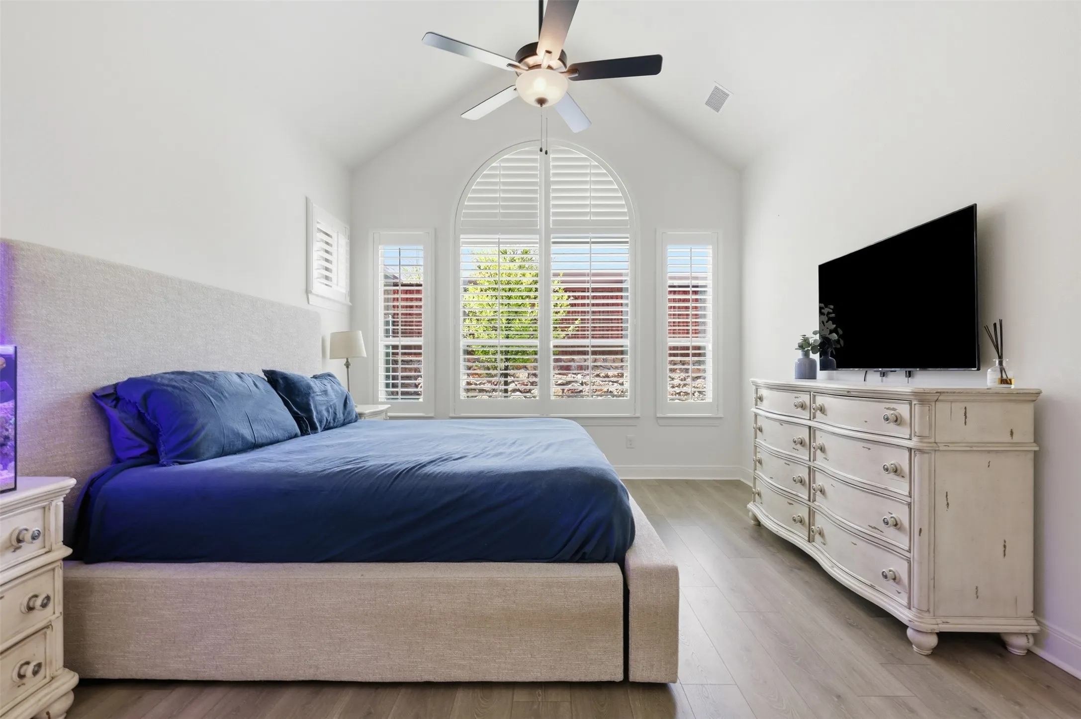 Vaulted ceiling with an integrated ceiling fan, arched windows featuring plantation shutters, wood-finish flooring, and crisp white walls