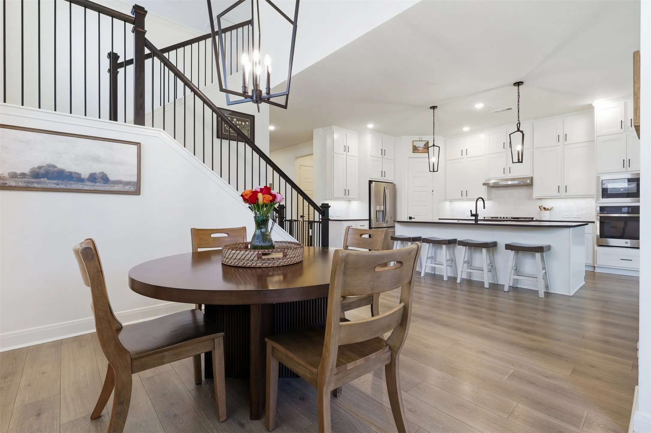 Open-concept living space featuring wood-finish flooring, a contemporary kitchen with white cabinetry and dark countertops, and a staircase with dark wood handrails and black metal balusters