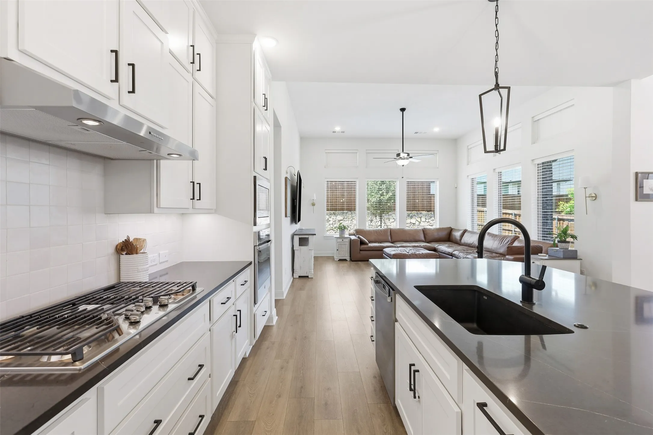 Open-concept kitchen with white cabinetry, dark countertops, a gas range, a central island with an undermount sink and gooseneck faucet, and wood-finish flooring