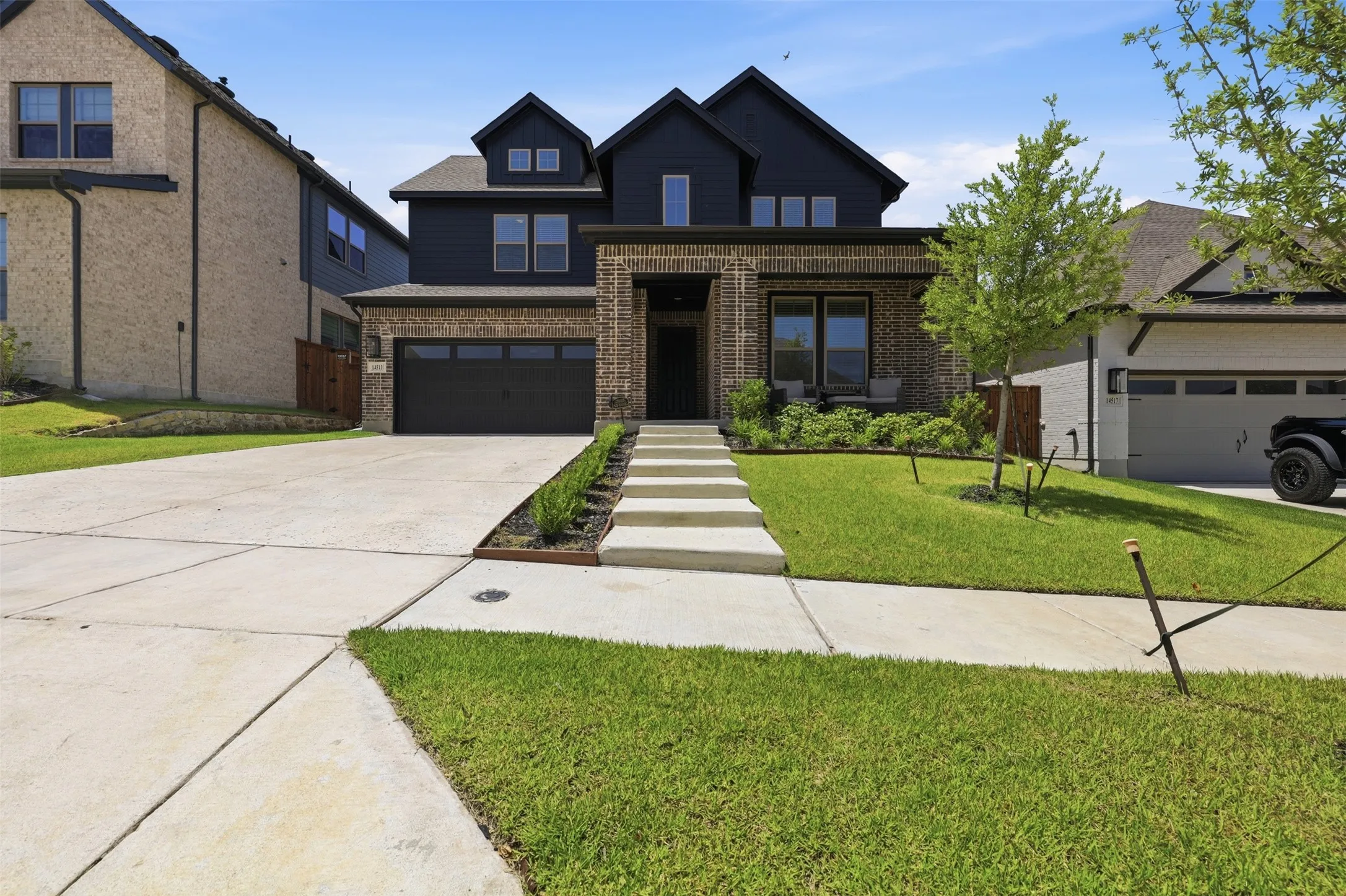 Modern two-story residence with a dark facade and brick accents, featuring a two-car garage, a concrete driveway, and a stone walkway with integrated landscaping