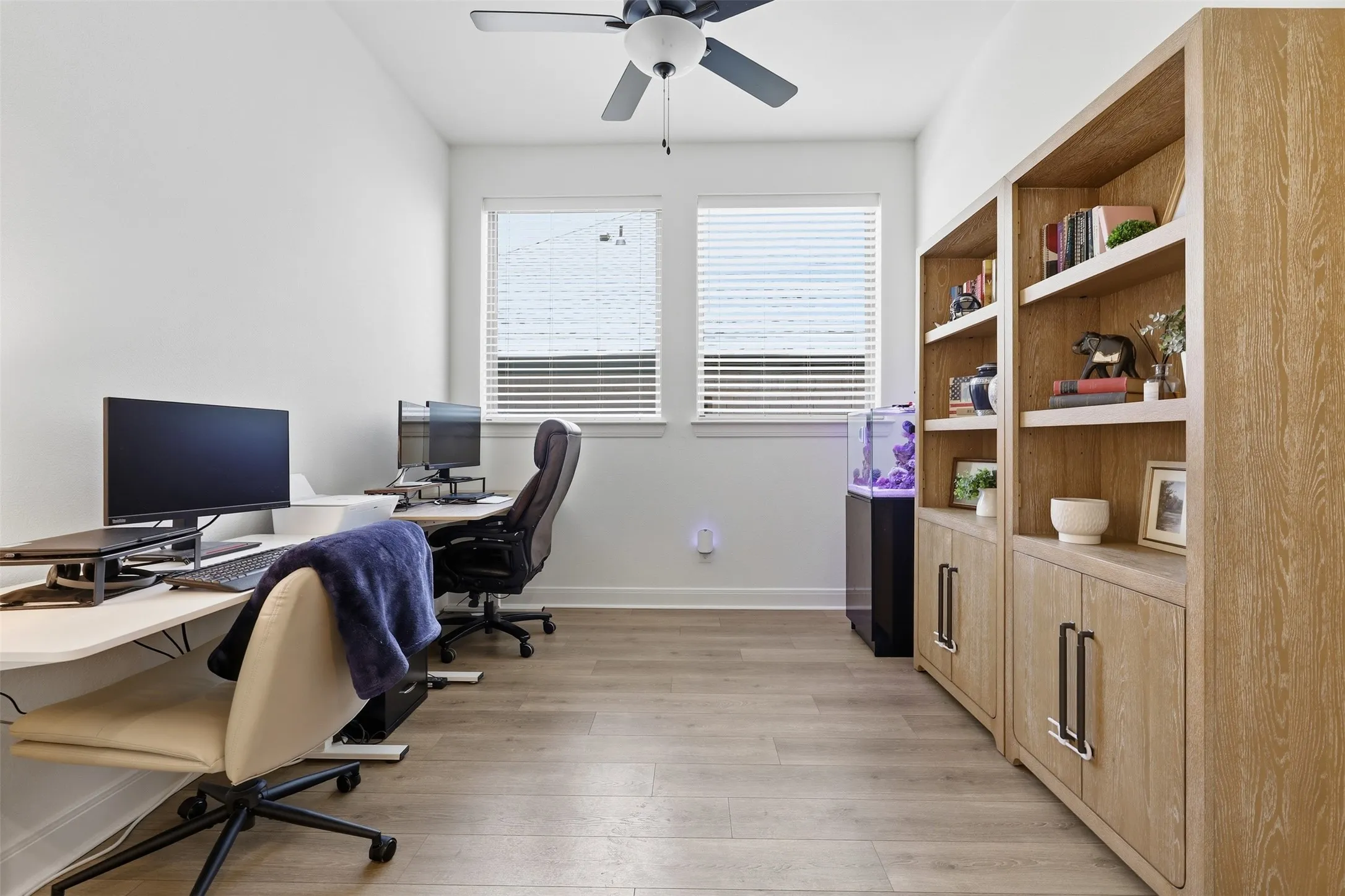 Well-lit interior space featuring wood-finish flooring, a ceiling fan, dual windows with blinds, and built-in shelving with lower cabinetry