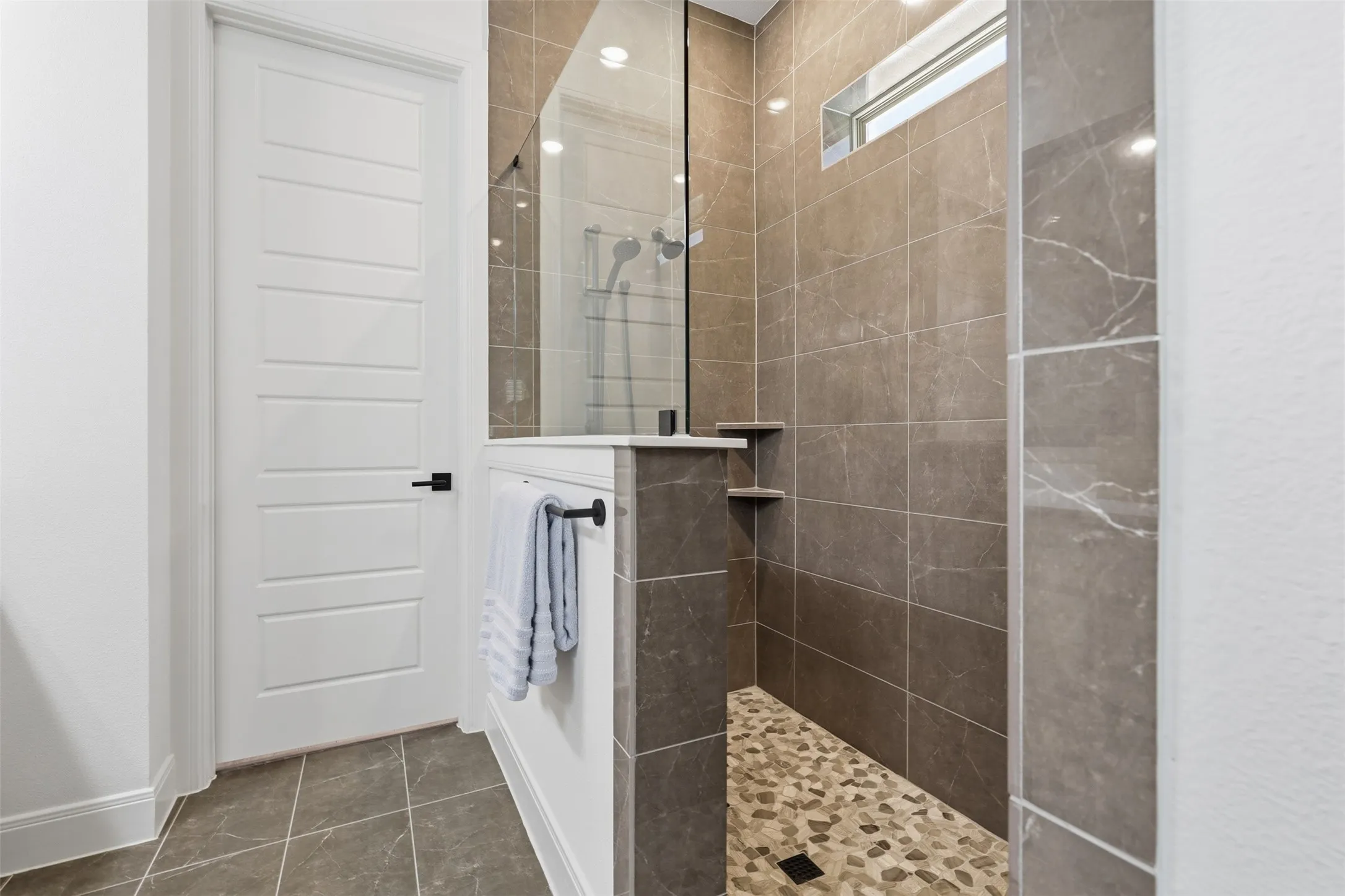Walk-in shower featuring glass enclosure, stone tile walls, pebble flooring, recessed lighting, and built-in shelving