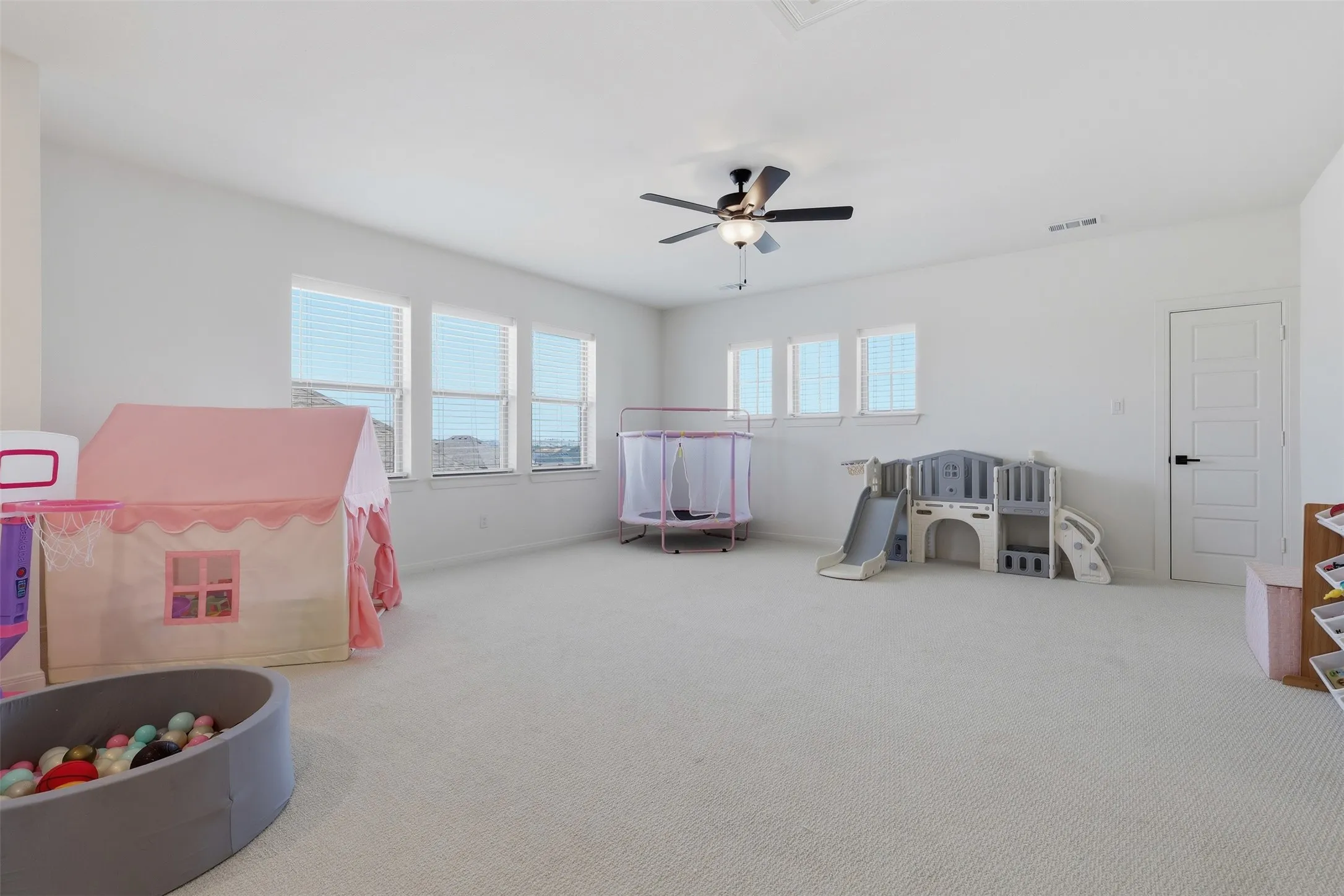 Spacious room featuring multiple windows with blinds, light-toned carpeting, a ceiling fan with integrated lighting, and a paneled interior door with matte black hardware