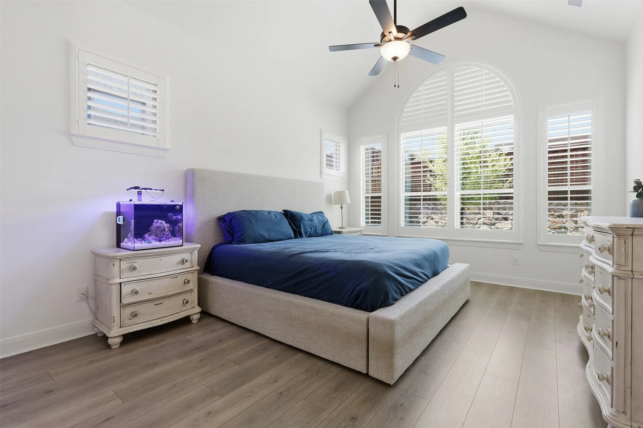 Room featuring a vaulted ceiling with a ceiling fan, wood-finish flooring, and multiple windows with white plantation shutters, including a large arched window