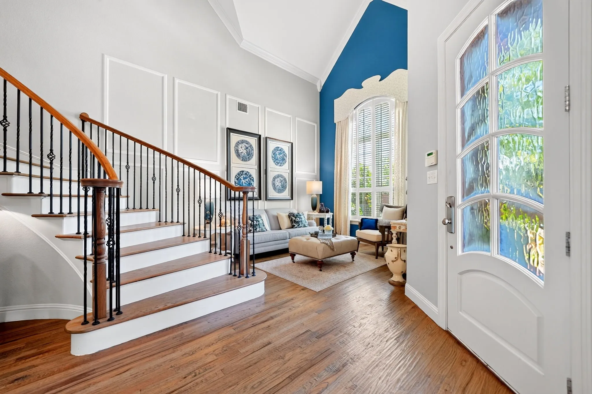 Entryway featuring wood-finish flooring, a white paneled door with leaded glass, a curved staircase with wood handrails and wrought iron balusters, high ceilings, and a living area with a large arched window