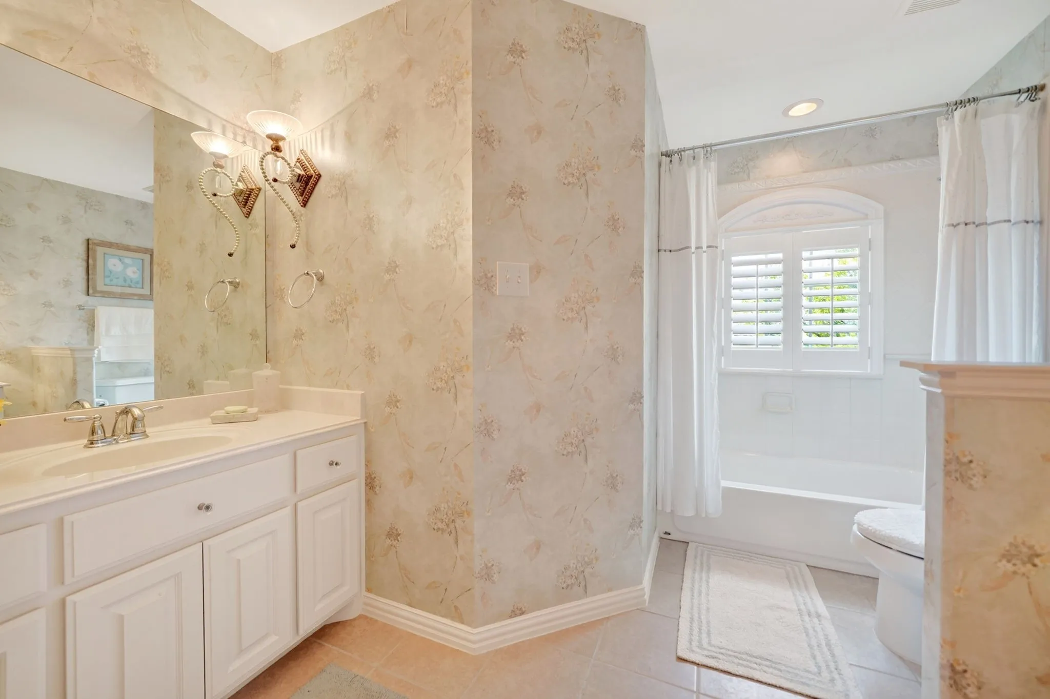 Bathroom featuring a white vanity with an integrated sink, an arched window with plantation shutters, a bathtub, a toilet, and patterned wallpaper