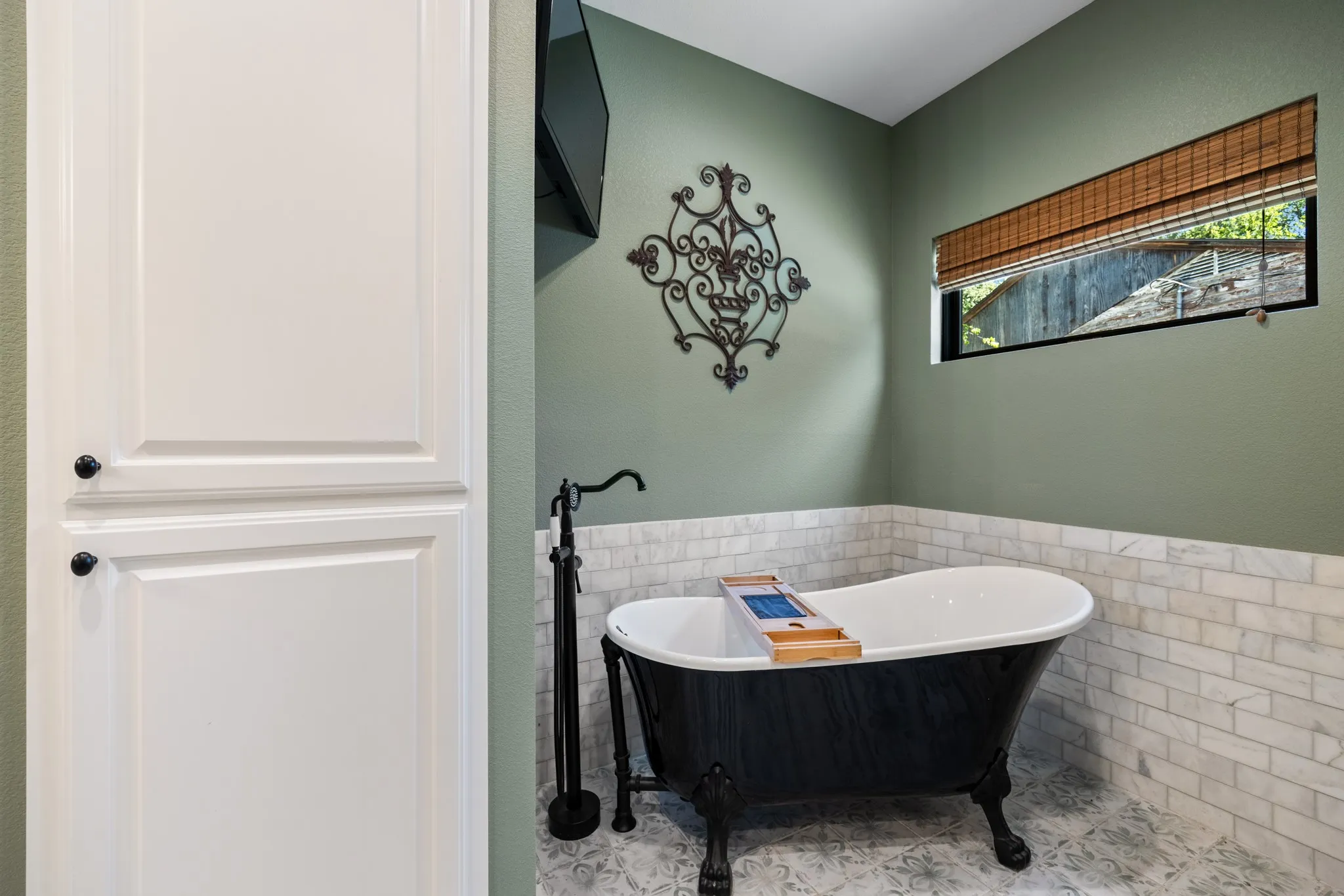 Freestanding clawfoot tub with a matte black floor-mounted faucet, white shaker-style built-in cabinetry, and white subway tile wainscoting