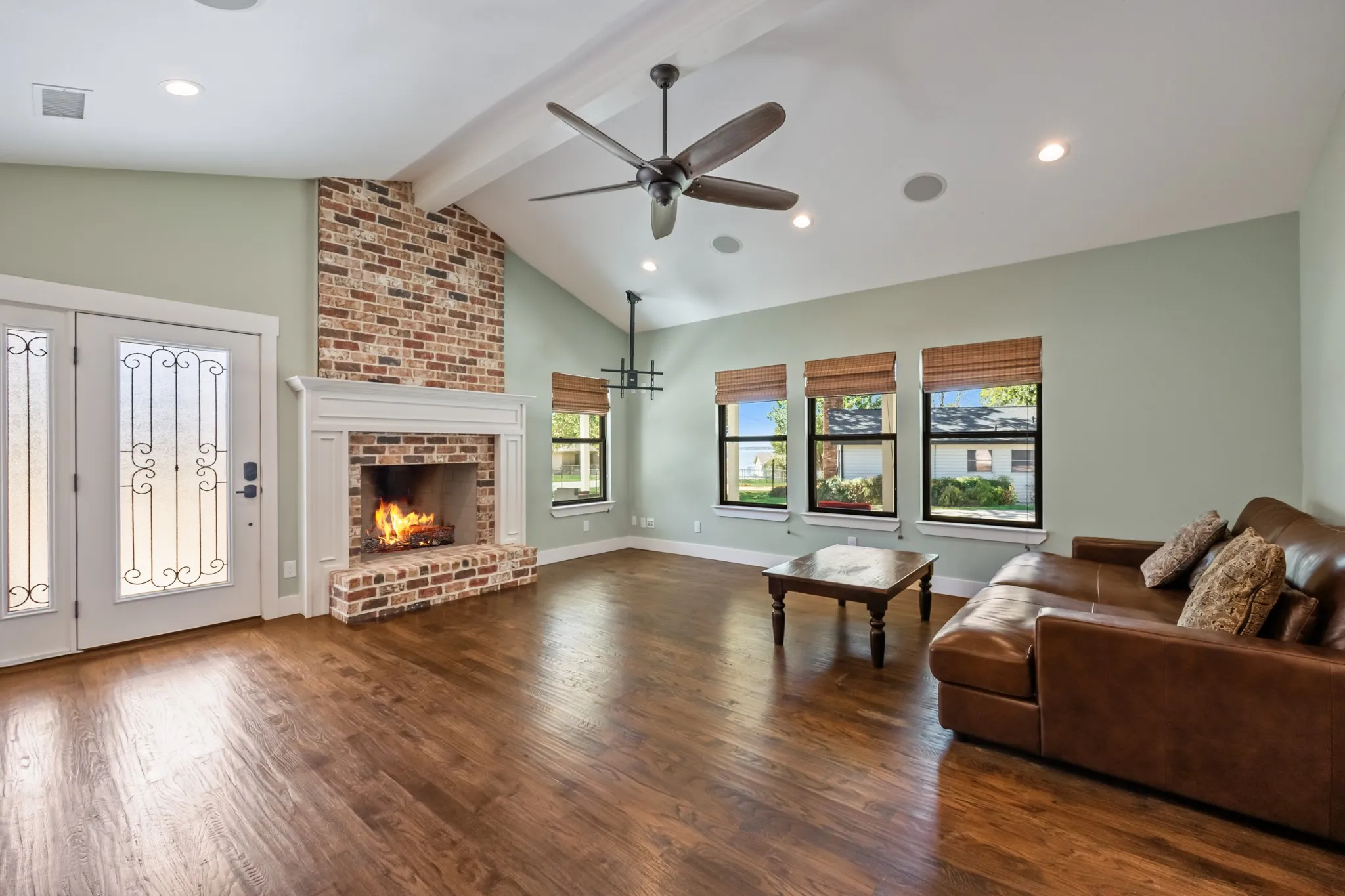 Vaulted ceiling with exposed beam, brick fireplace with mantel, dark wood-finish flooring, recessed lighting, and multiple windows