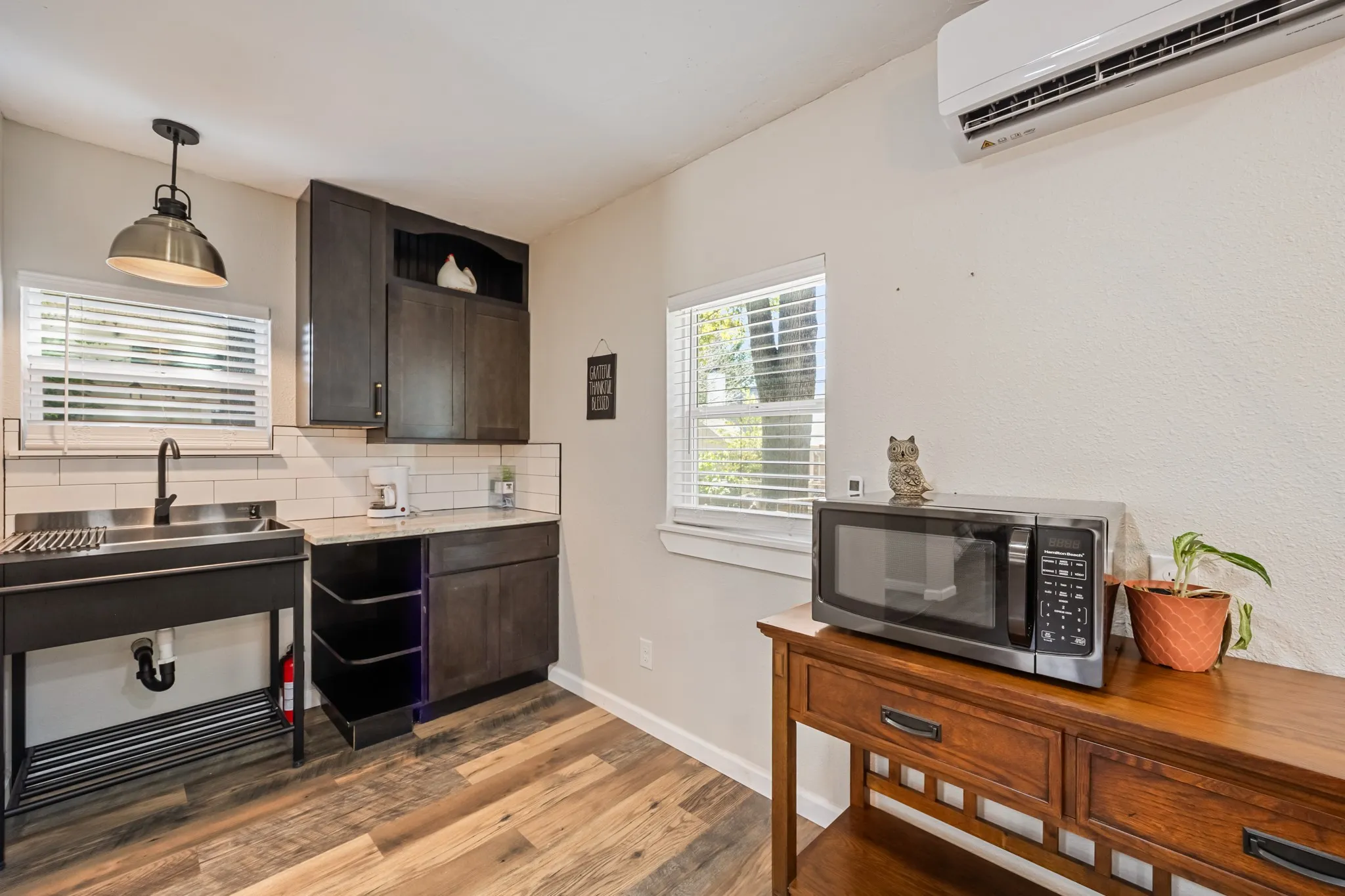 Compact kitchen area featuring wood-finish flooring, dark wood-tone cabinetry, white subway tile backsplash, a stainless steel sink, and a wall-mounted mini-split unit