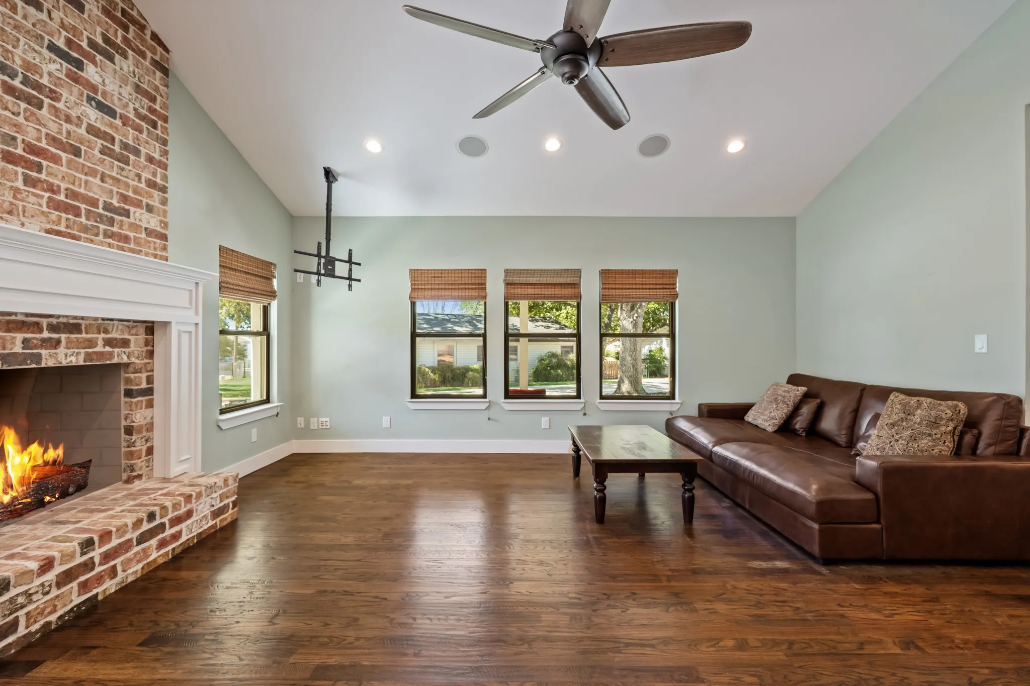 Vaulted ceiling with a ceiling fan, recessed lighting, brick fireplace with a white mantel, three windows, and dark wood-finish flooring