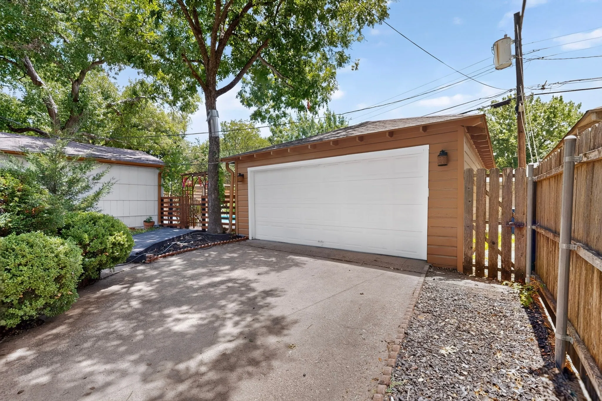 Detached garage with a white overhead door, brown siding, and a side-gate entrance, bordered by a gravel path and mature trees