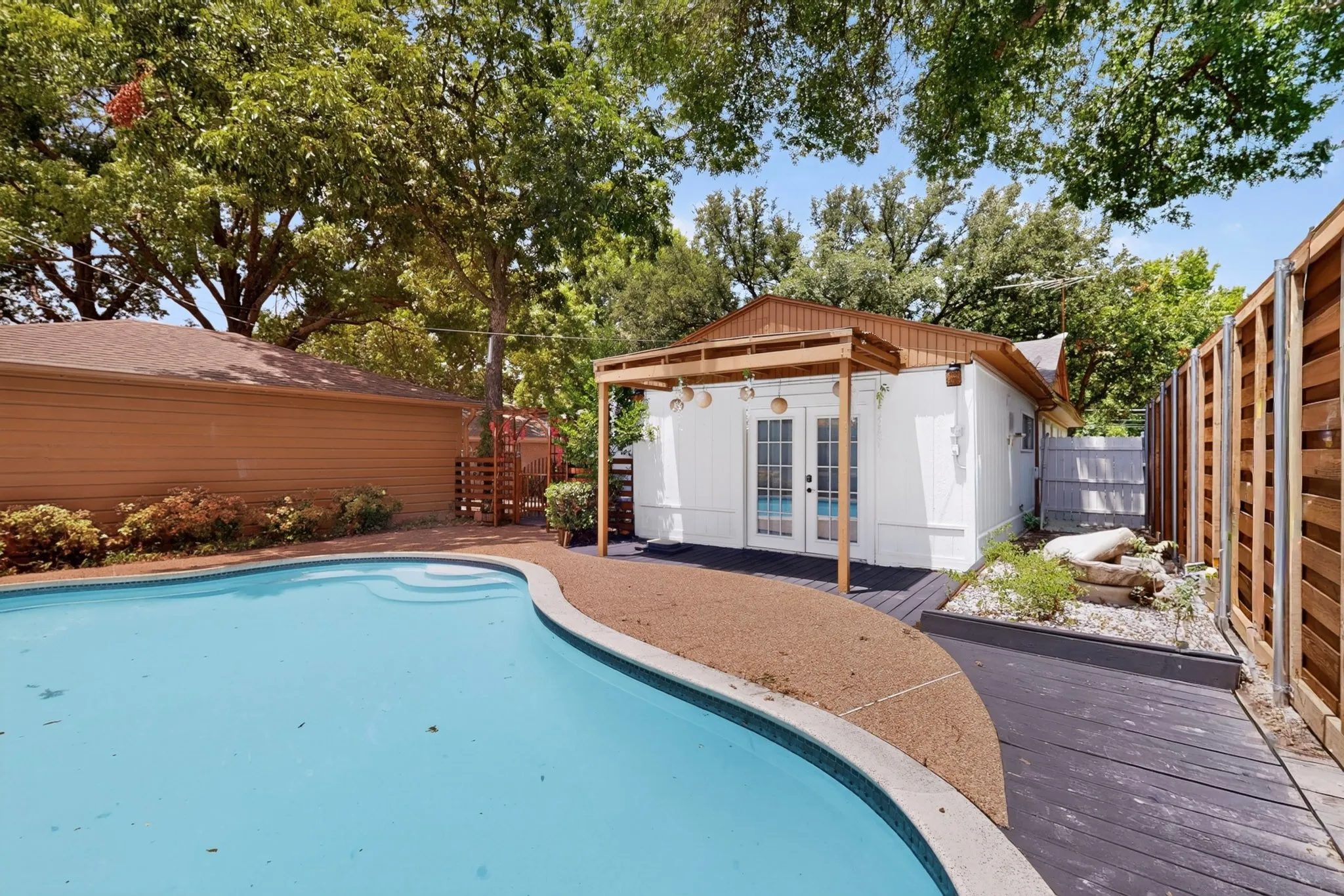 Curved swimming pool with integrated steps, complemented by an adjacent white auxiliary building featuring French doors and a wood pergola