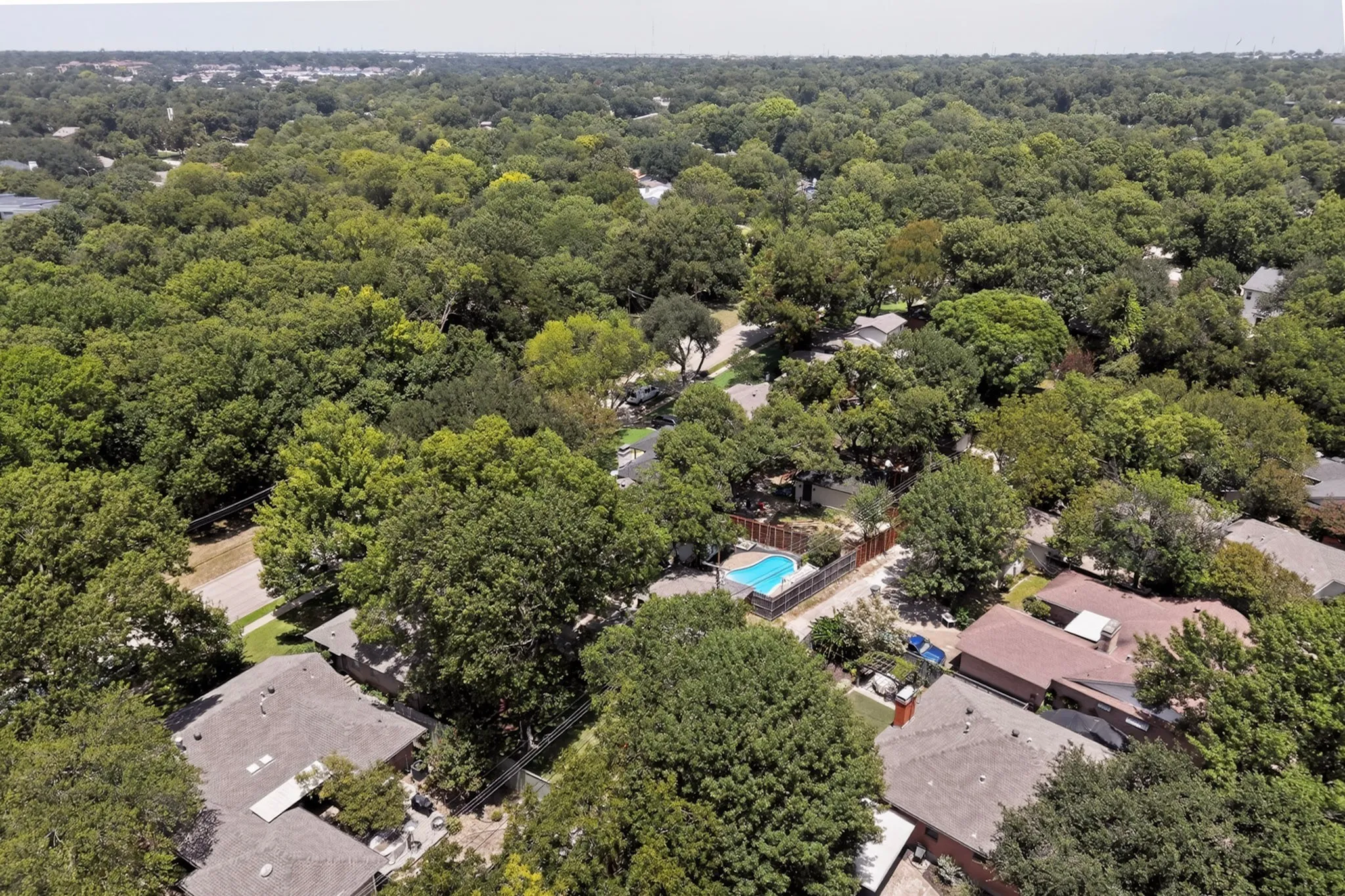 Aerial perspective of a verdant residential area featuring numerous mature trees, individual homes with shingle roofs, and a backyard swimming pool