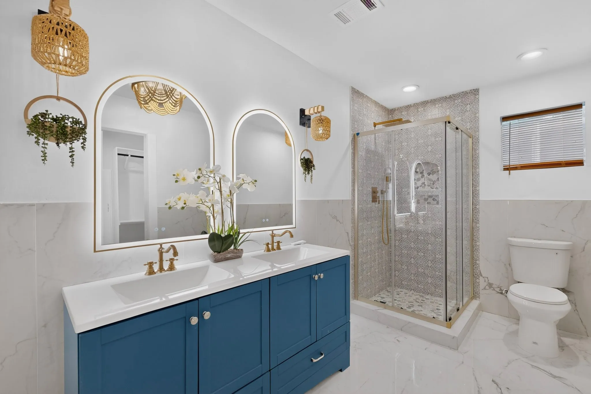 Full bathroom featuring a double vanity with a blue finish, dual arched backlit mirrors, a glass-enclosed shower with tiled walls, and white and gray marble-finish flooring and wainscoting