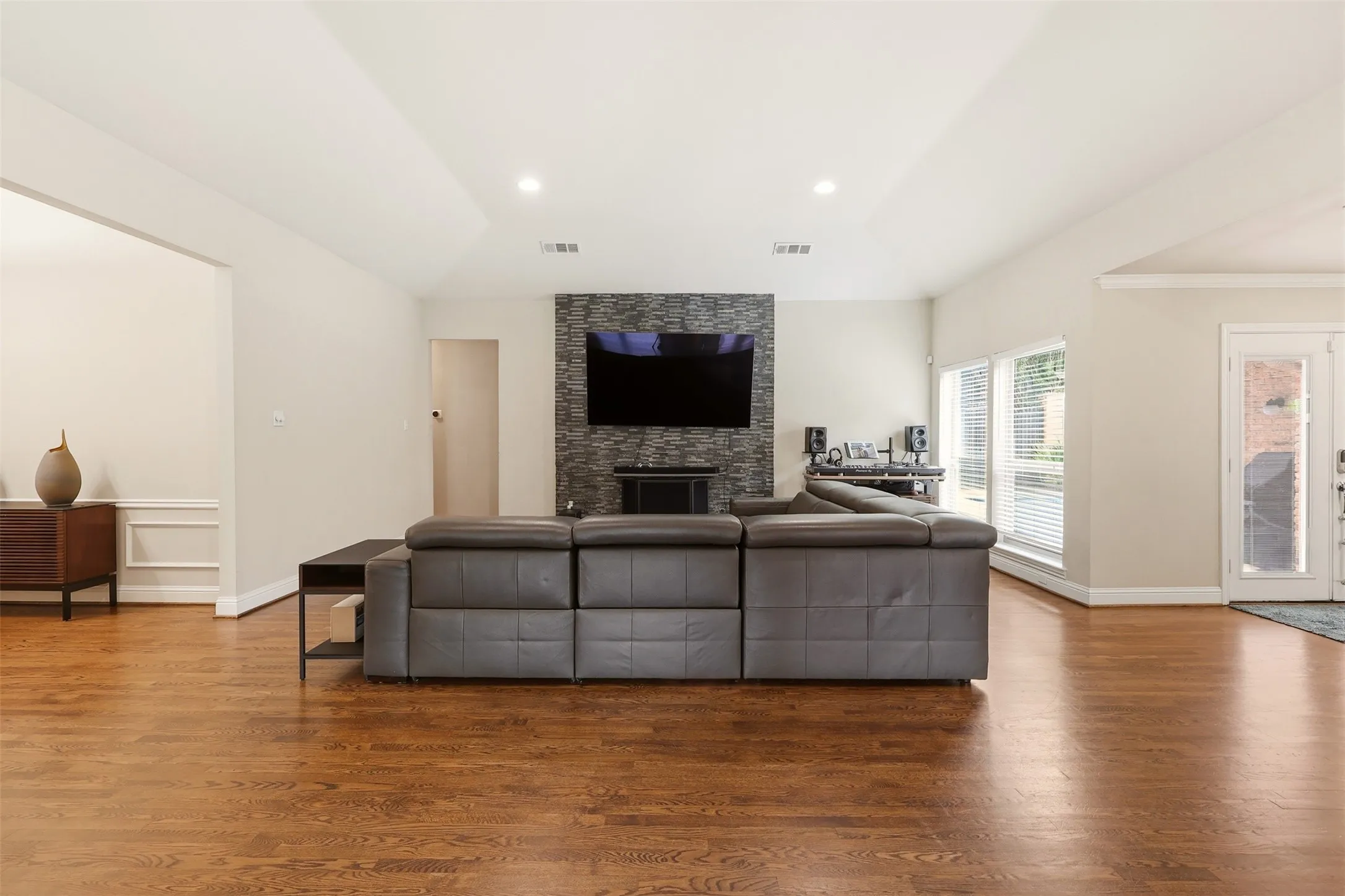 Spacious living area featuring wood-finish flooring, a vaulted ceiling with recessed lighting, and a stone-clad accent wall