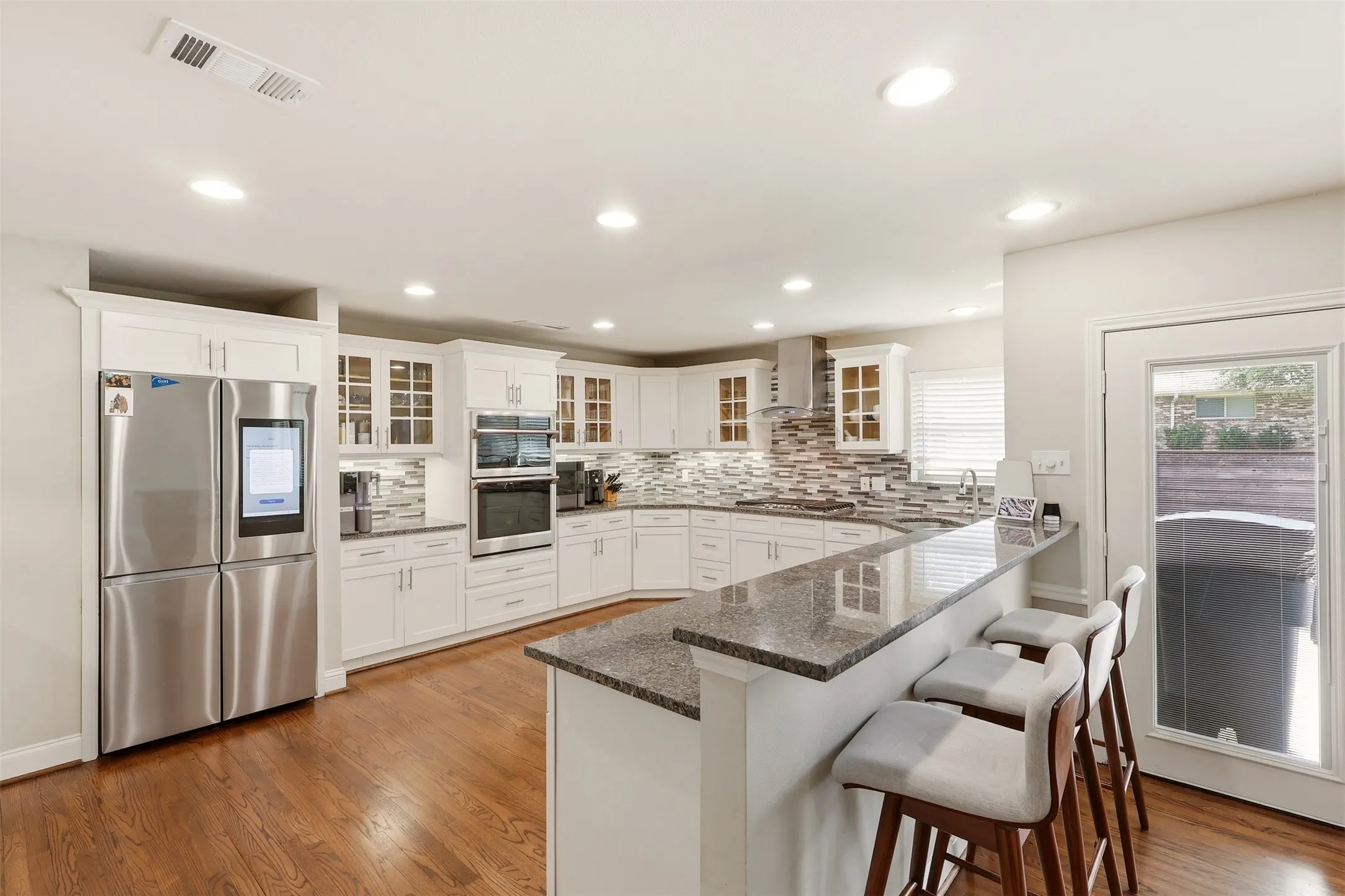 Bright kitchen featuring recessed lighting, extensive white cabinetry, stainless steel appliances, a multi-tone tile backsplash, and a granite-topped peninsula with bar seating