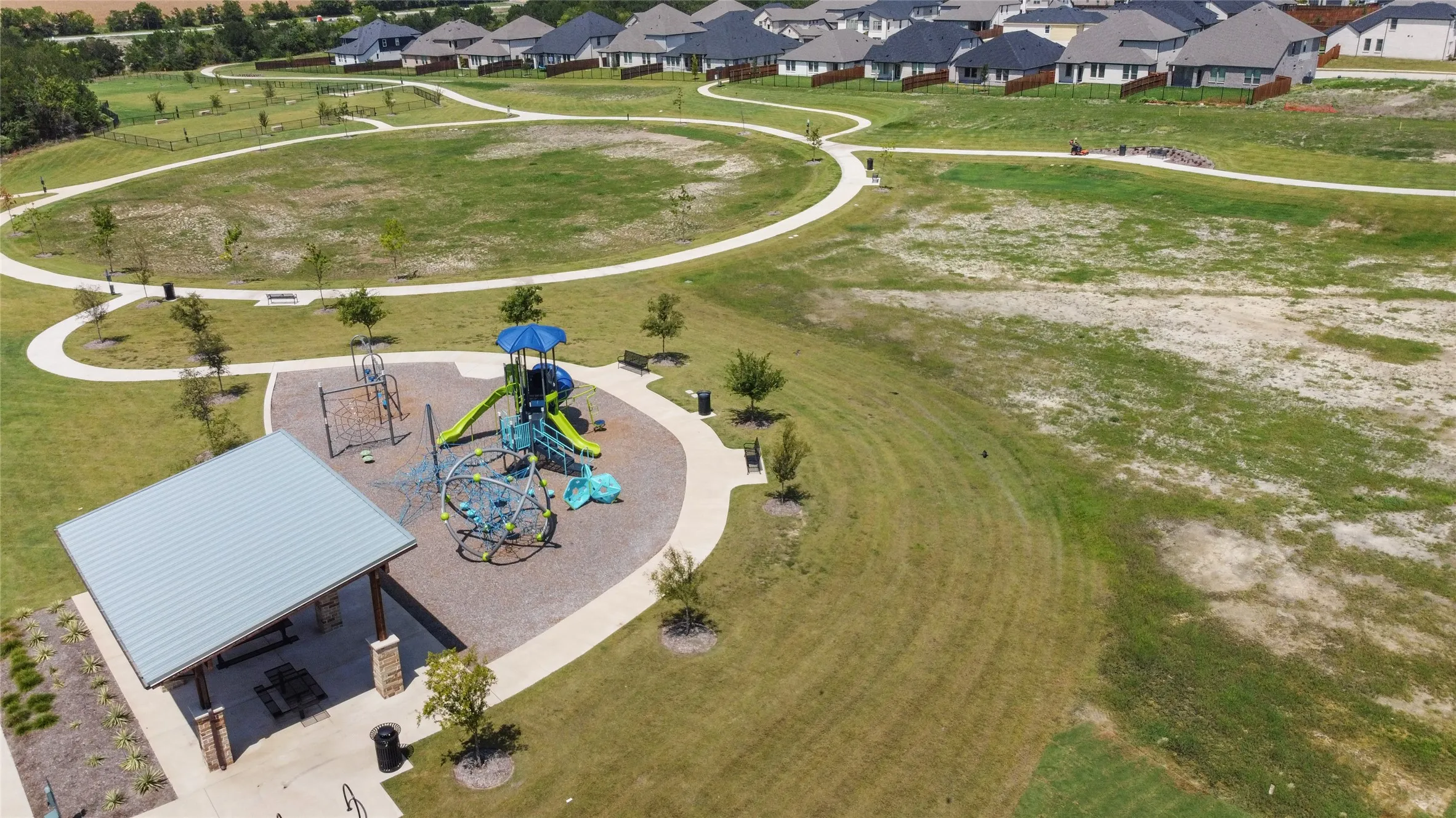 Community park featuring a covered picnic area, a large playground with multiple slides and climbing structures, paved walking paths, and open grassy areas