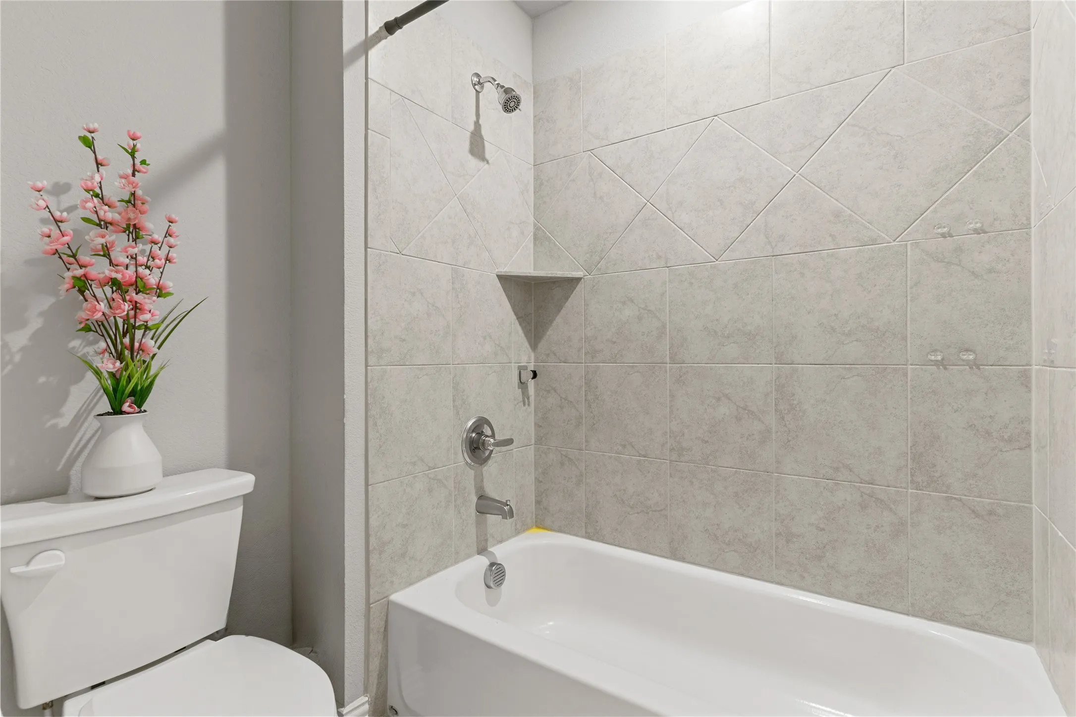 Bathtub with floor-to-ceiling gray tile surround, featuring a built-in corner shelf and polished chrome fixtures