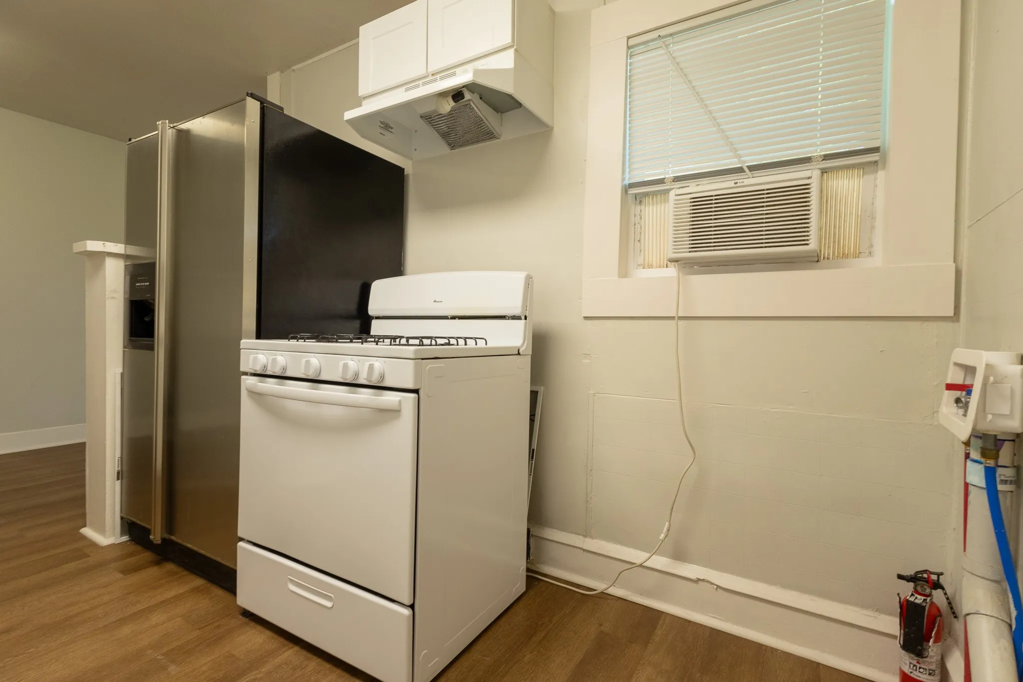 Kitchen area featuring wood-finish flooring, a white gas range with overhead exhaust, stainless steel refrigerator with an ice/water dispenser, and a window with an integrated air conditioning unit