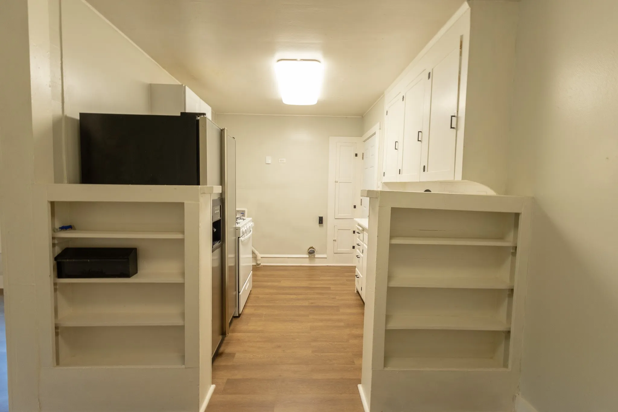 Galley kitchen featuring wood-finish flooring, white built-in shelving, stainless steel refrigerator, white range, and white cabinetry