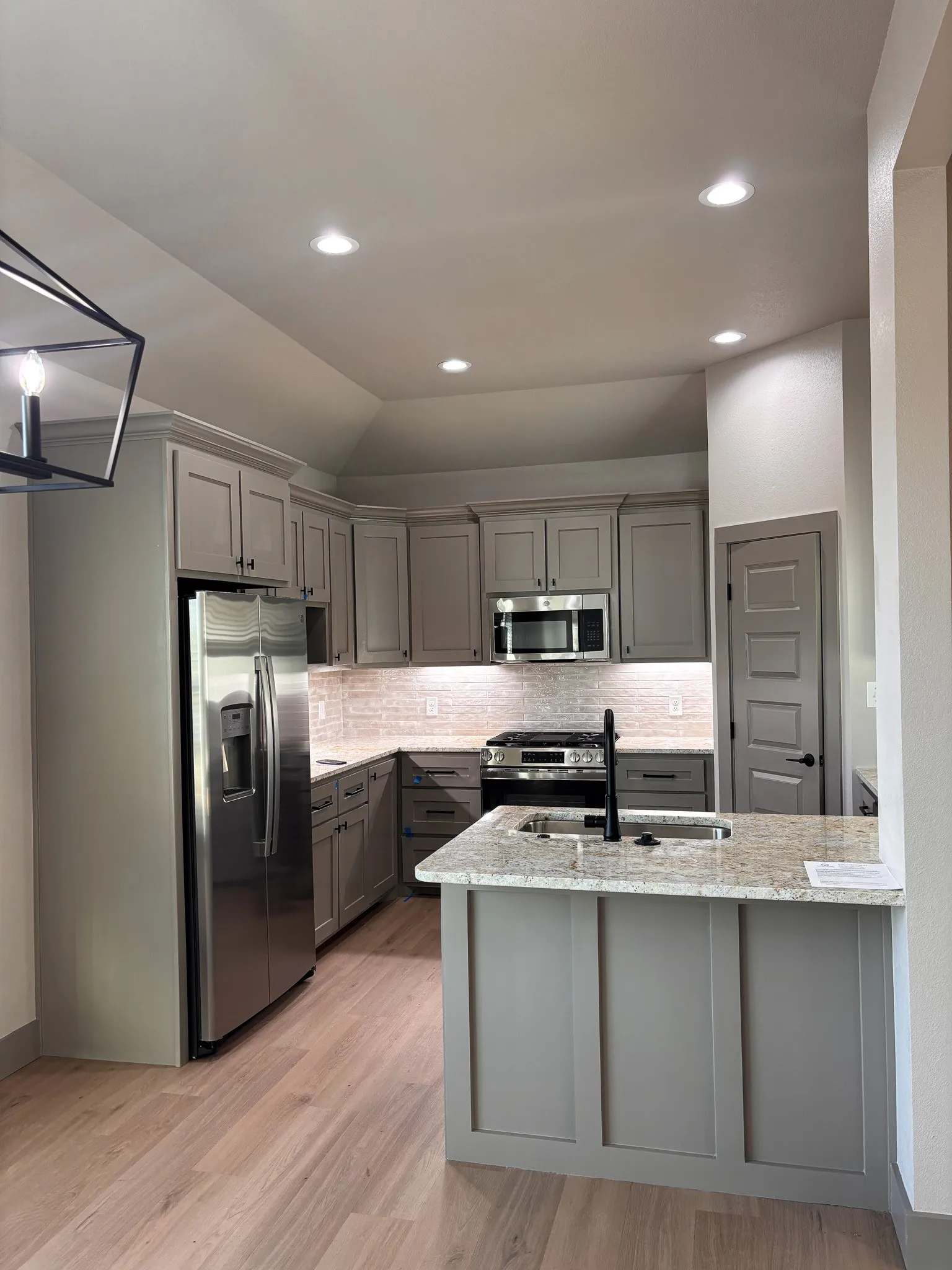 Modern kitchen featuring light wood-finish flooring, recessed lighting, stainless steel appliances, a light-toned backsplash, and a central island with a sink and dark faucet