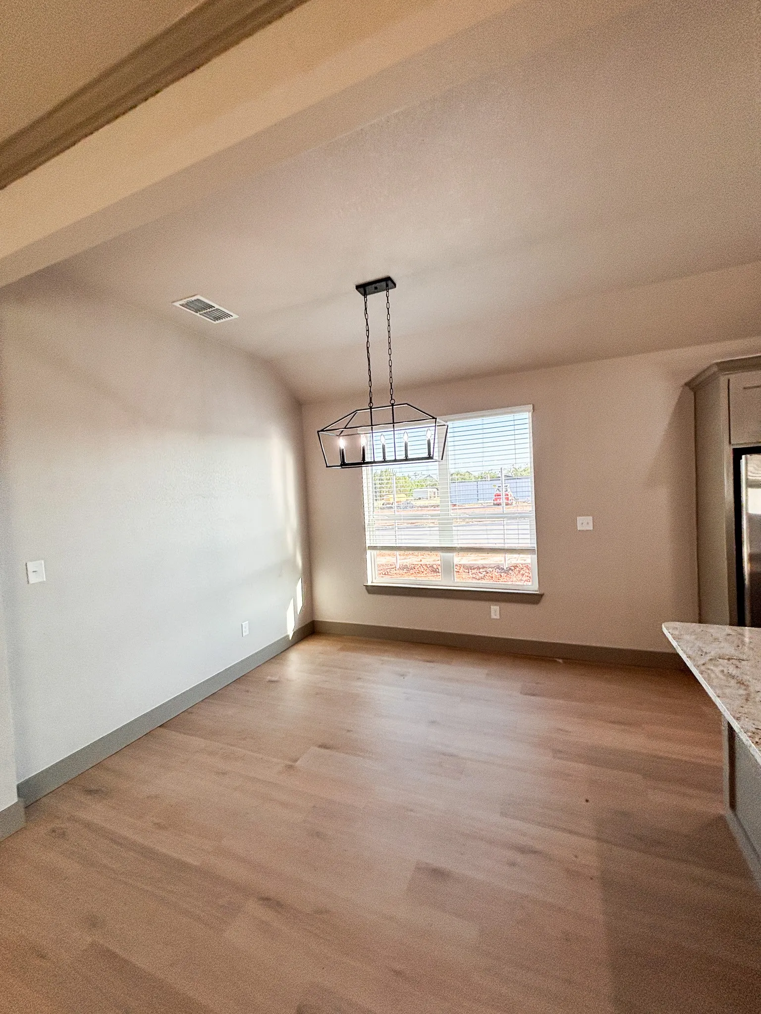 Spacious room featuring wood-finish flooring, a contemporary black geometric chandelier, and a large window with blinds