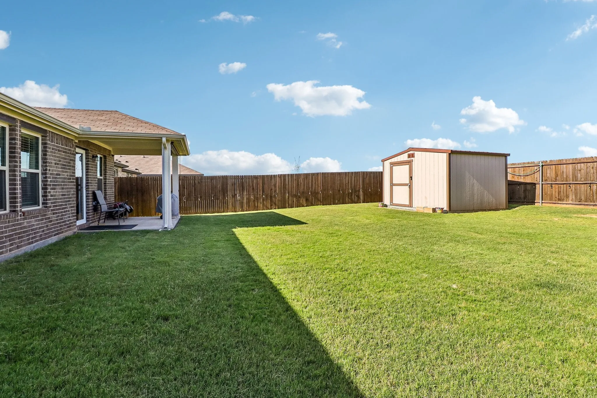 Expansive green lawn with a covered patio, brick exterior, wood privacy fence, and a detached storage shed