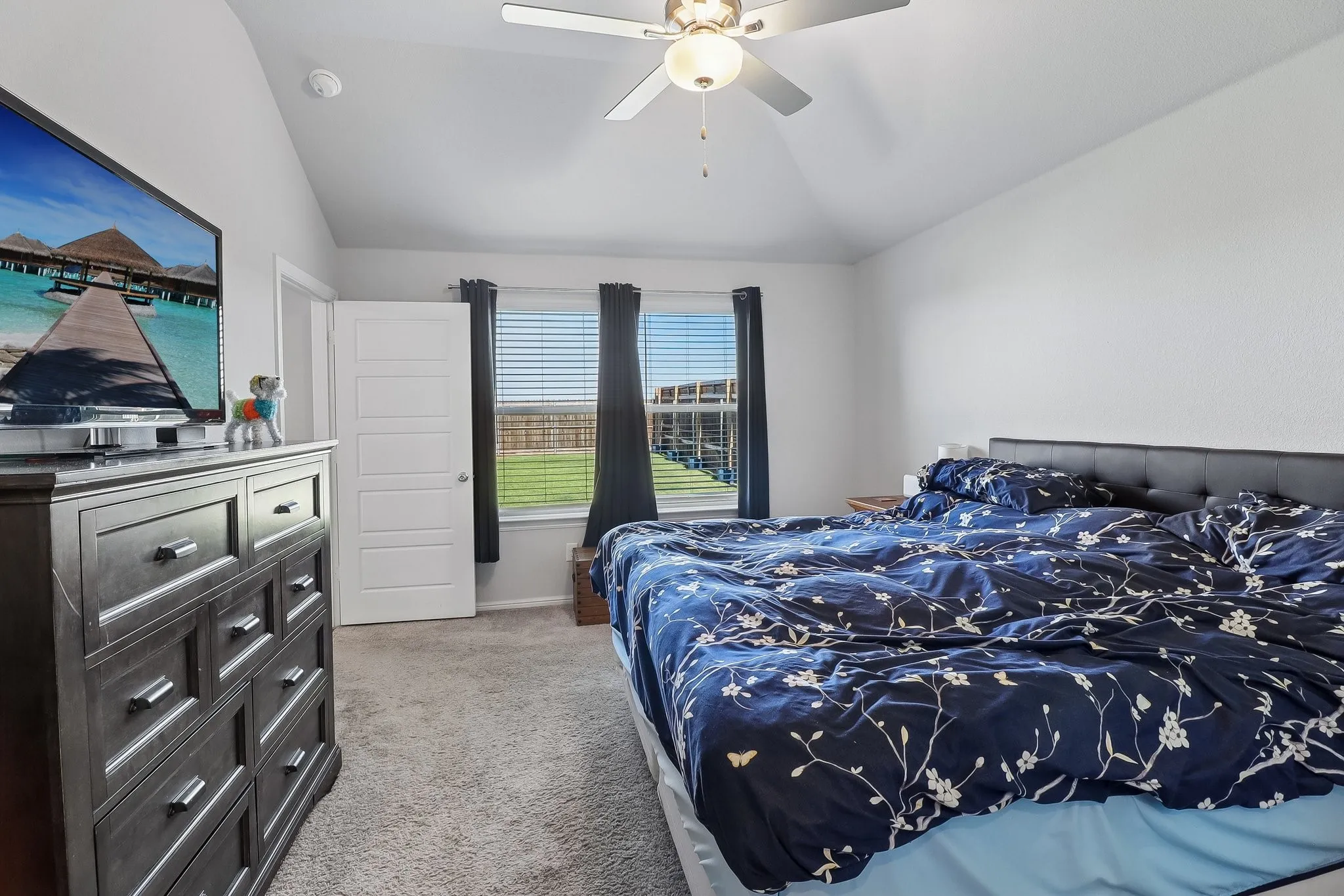 Spacious room featuring vaulted ceilings, a ceiling fan with integrated lighting, carpeted flooring, and a pair of windows with horizontal blinds