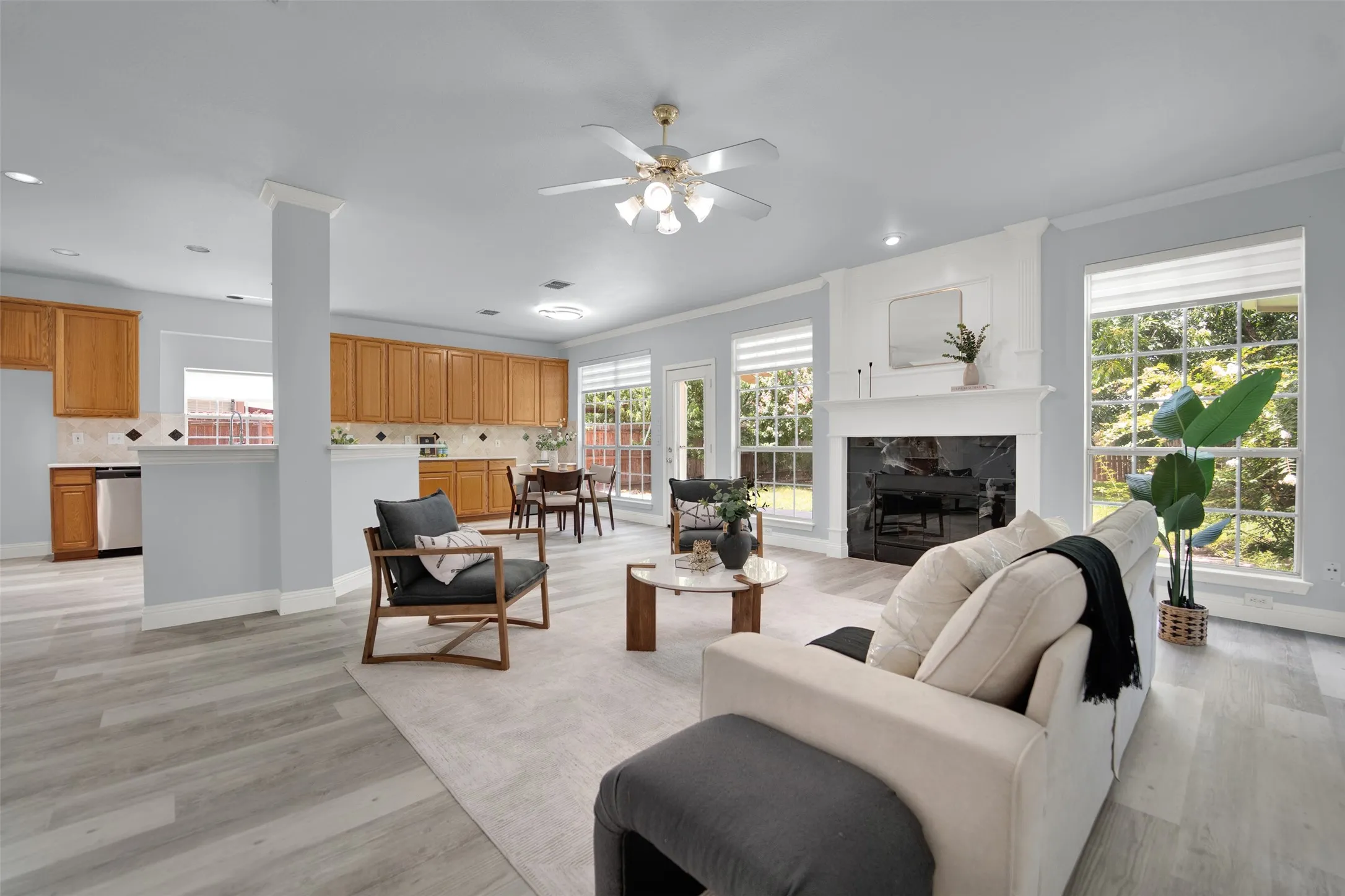 Spacious living area featuring light wood-finish flooring, a white mantel fireplace with a dark stone surround, and multiple large windows