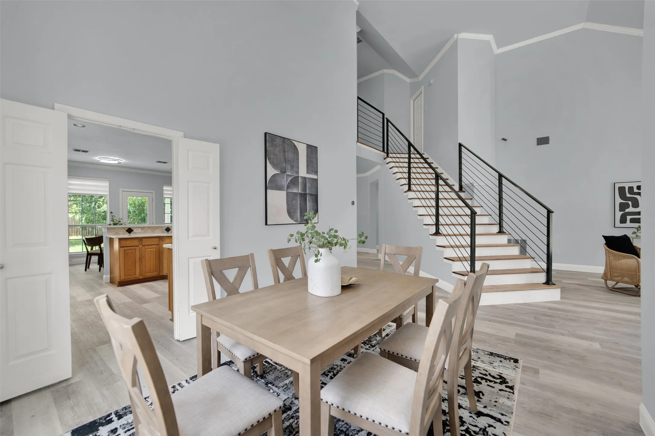 Dining area featuring light-toned wood-finish flooring, a contemporary staircase with black metal railings, and a double doorway leading to a kitchen with wood cabinetry and tile countertops