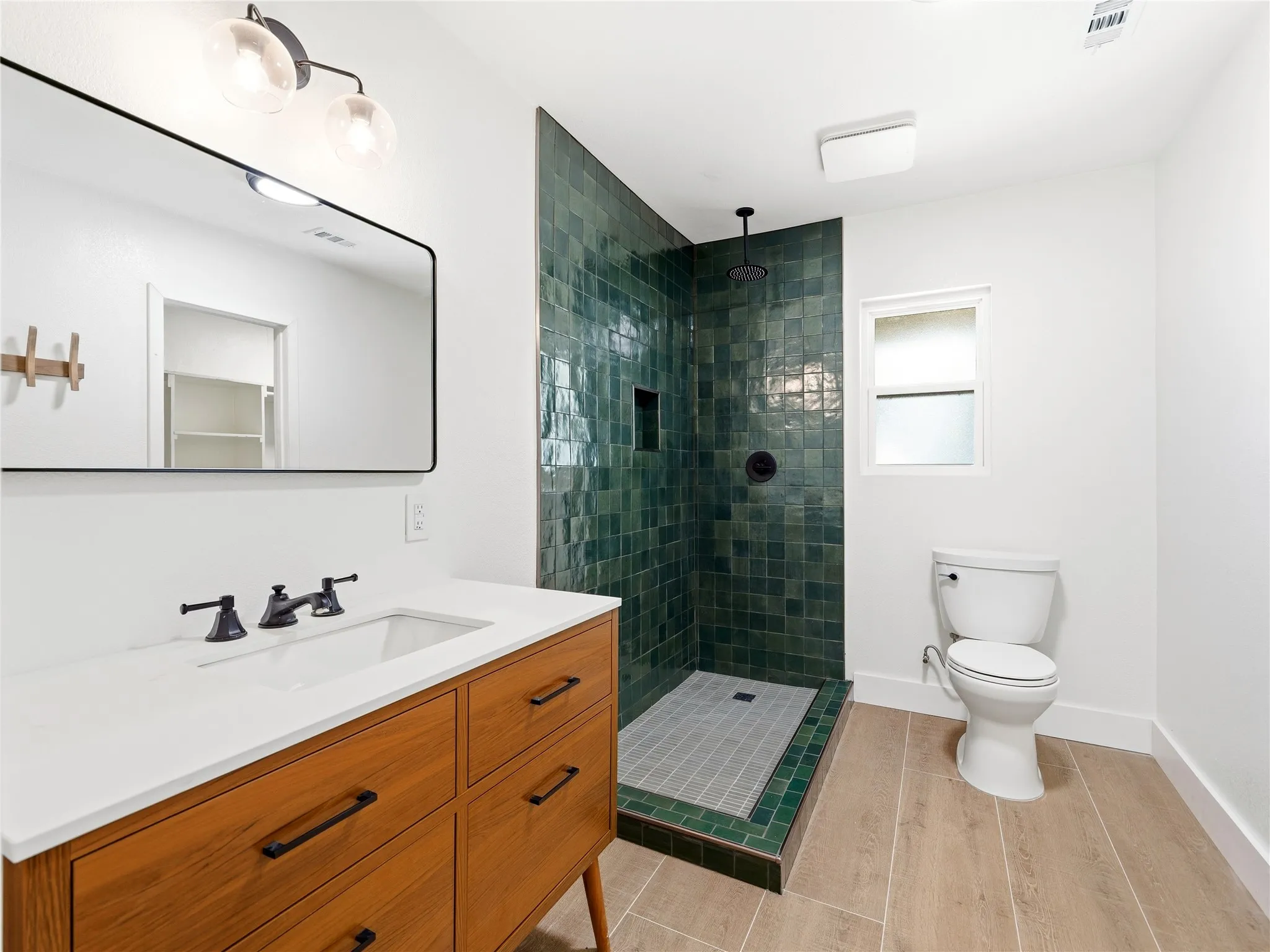 Bathroom featuring a wood-finish vanity with a white countertop and rectangular sink, matte black fixtures, a rectangular vanity mirror, a glass-enclosed shower with green tile surround, and wood-finish flooring